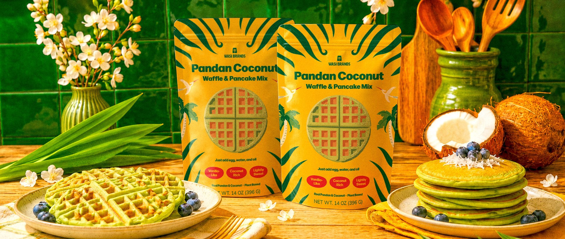 Two pouches of Wasi Brands Pandan Coconut Waffle & Pancake Mix displayed on a wooden countertop with green waffles, pancakes, pandan leaves, coconuts, and ceramic kitchenware.