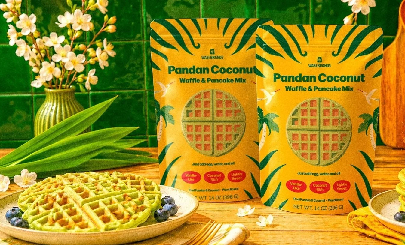 Wasi Brands Pandan Coconut Waffle & Pancake Mix packaging displayed in a styled kitchen setting with pandan leaves, green waffles, and floral accents.