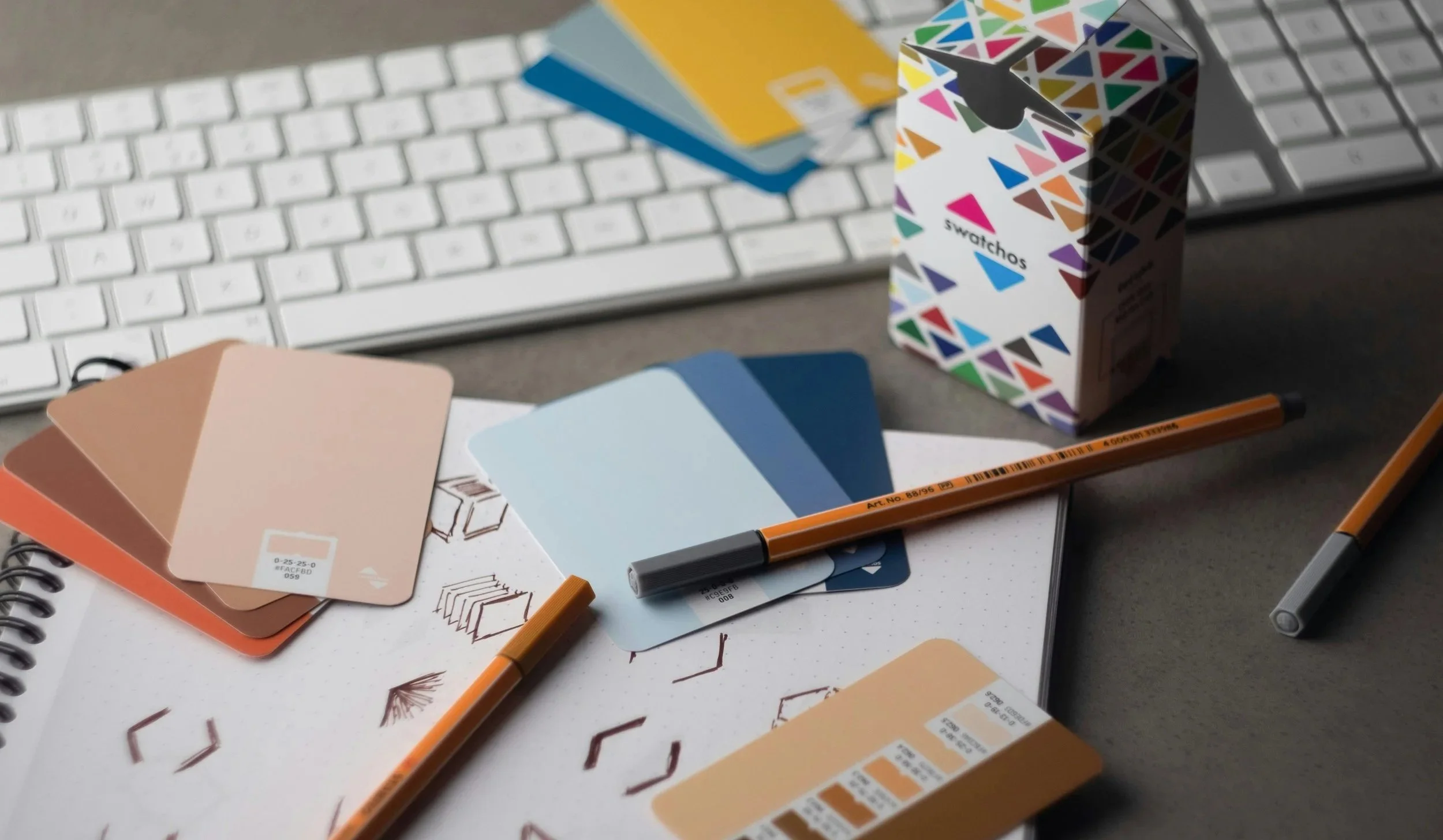 Color palette swatches, pens, a keyboard, lavender-colored card, and a colorful Swatchos box on a desk.