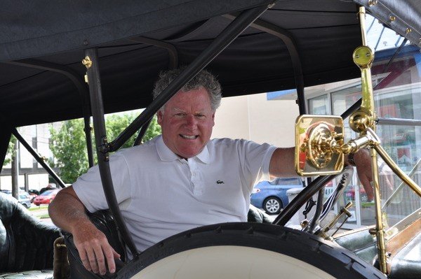 A man with curly gray hair sitting in a vintage car with a black roof, white body, and gold accents, smiling and pointing downward.