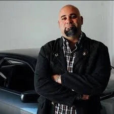 A smiling man with a shaved head, crossing his arms, wearing a dark blazer and matching shirt, standing in front of a white background.