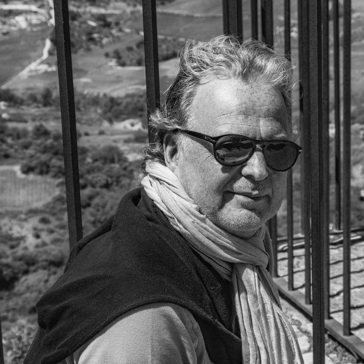 A man wearing sunglasses and a scarf, standing near a metal railing outdoors with a hilly landscape in the background.