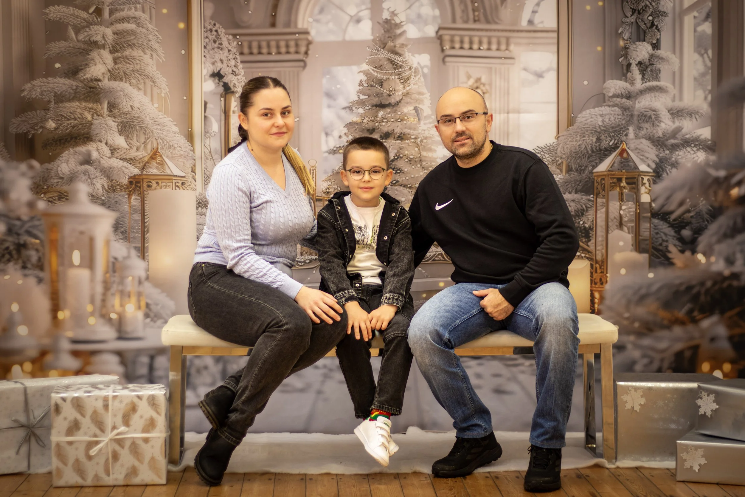 A family of four sitting on a bench in front of a holiday-themed winter backdrop with snow-covered trees, lanterns, and wrapped presents.