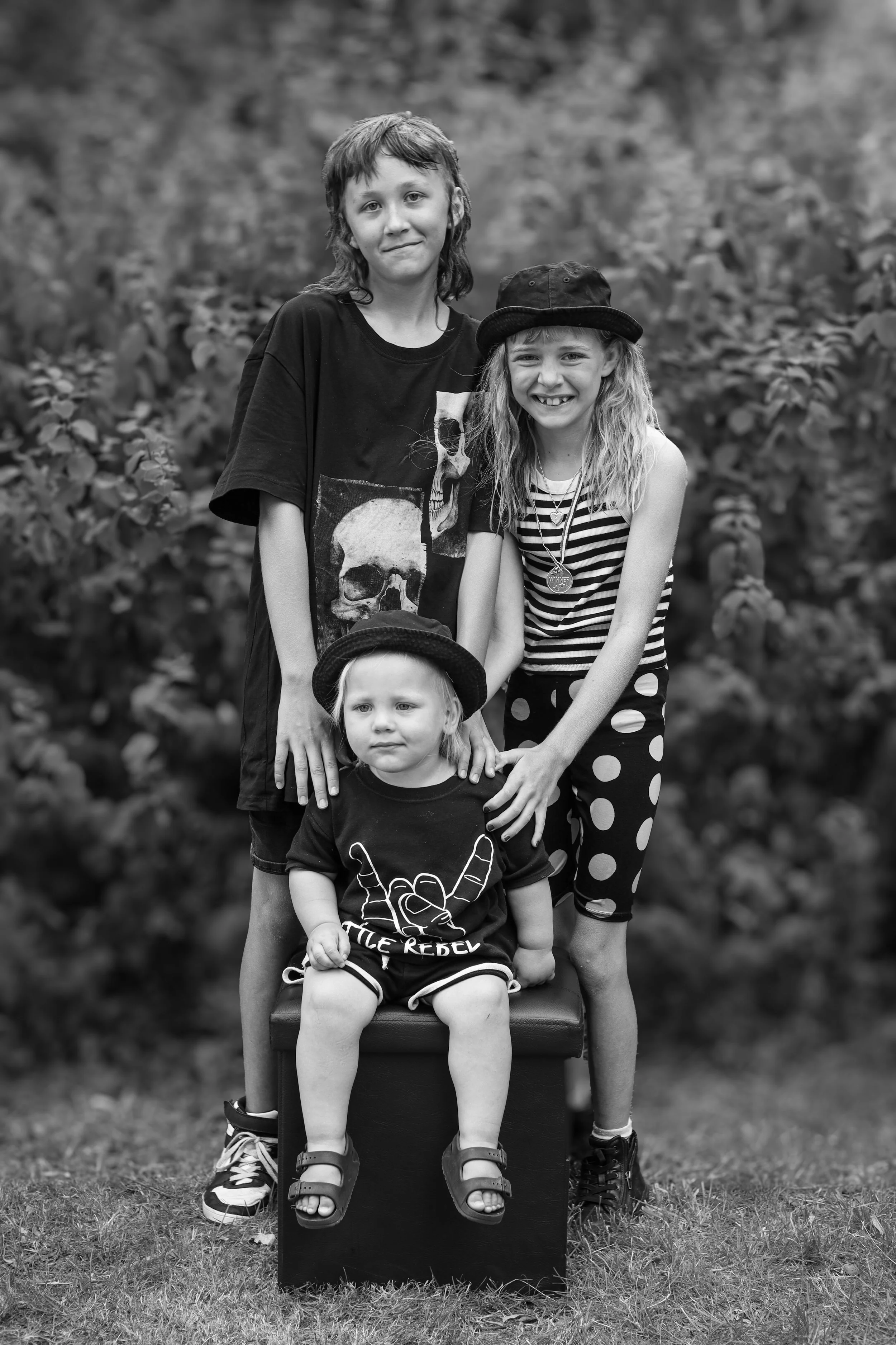 Three children, two girls and one boy, standing outdoors on grass with bushes in the background. The girl on the right wears a striped shirt and polka dot pants, smiling. The girl on the left is taller, with a skull graphic T-shirt, and has a neutral expression. The toddler girl seated on a box wears a black t-shirt with a hand making a rock and roll gesture and a hat, looking to the side.