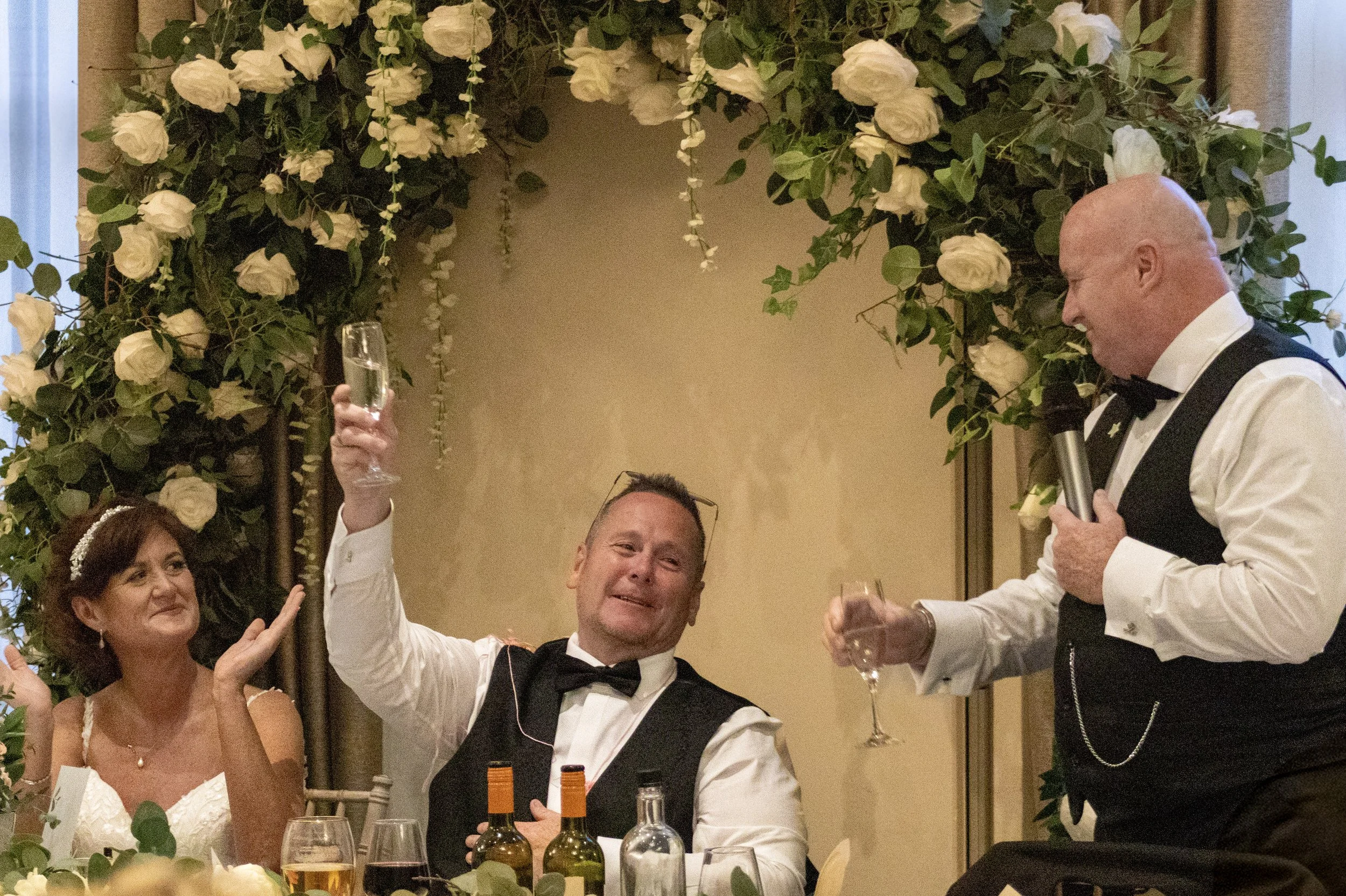 A group of people at a wedding reception, with two men in tuxedos raising glasses for a toast, one man smiling and the woman sitting beside them, all sitting at a decorated table with bottles and glasses, under a floral arch.