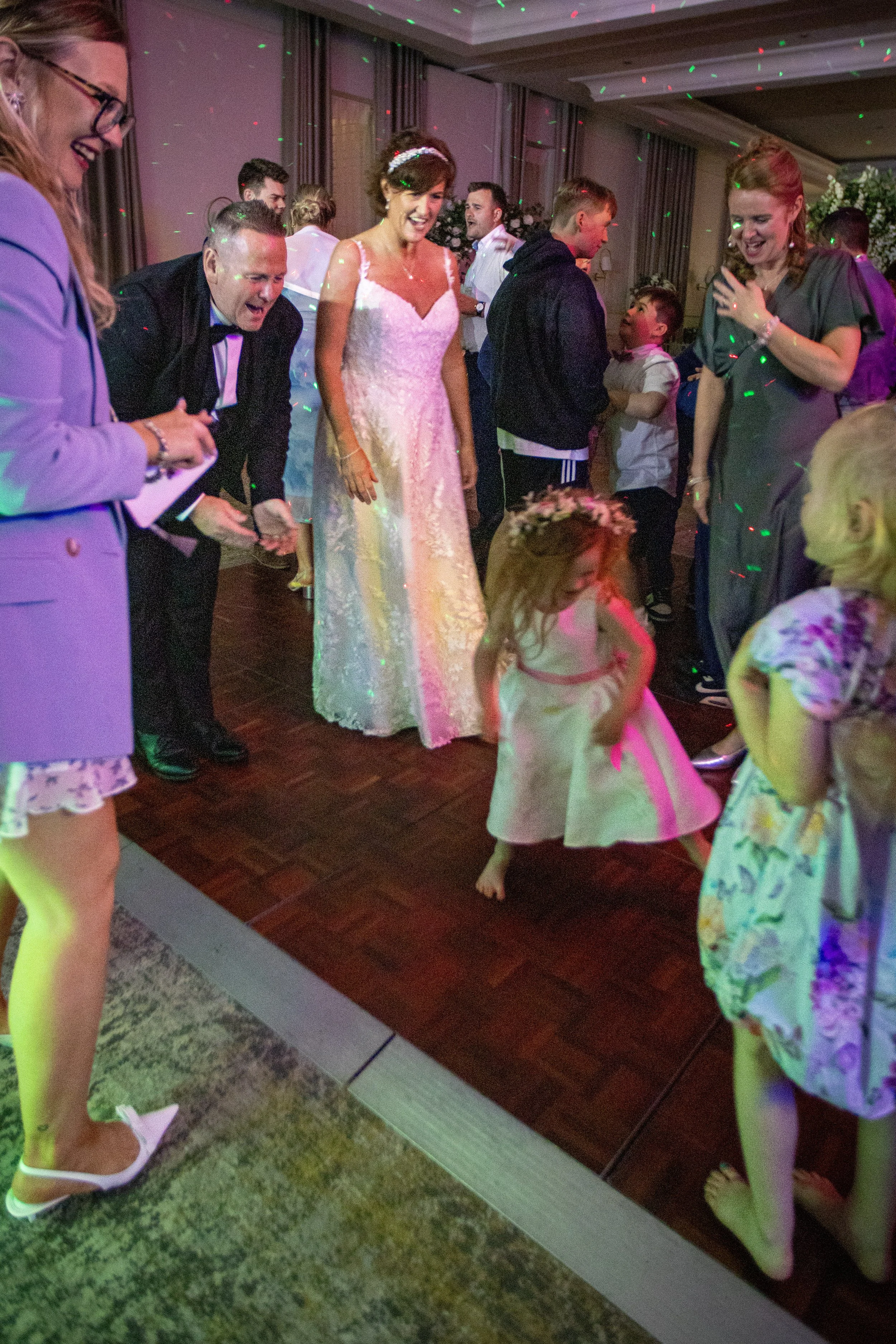 Group of people dancing and celebrating at a wedding reception, with a bride in a white wedding dress at the center, and children and adults joyfully dancing on the dance floor.