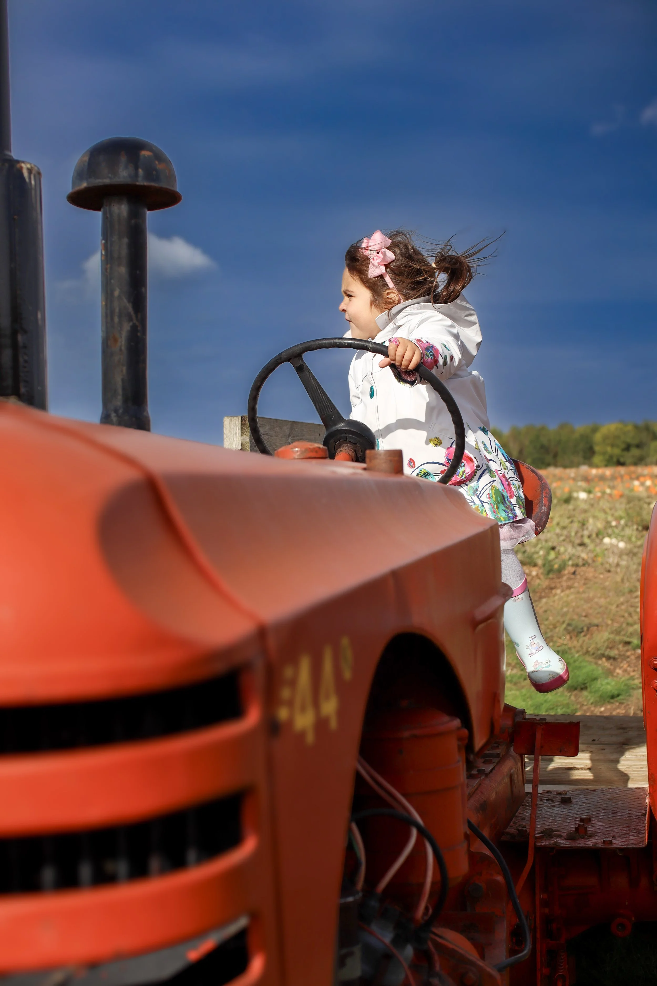 A young girl with a pink bow in her hair, wearing a white coat and colorful dress, sitting on an orange tractor outdoors under a blue sky.