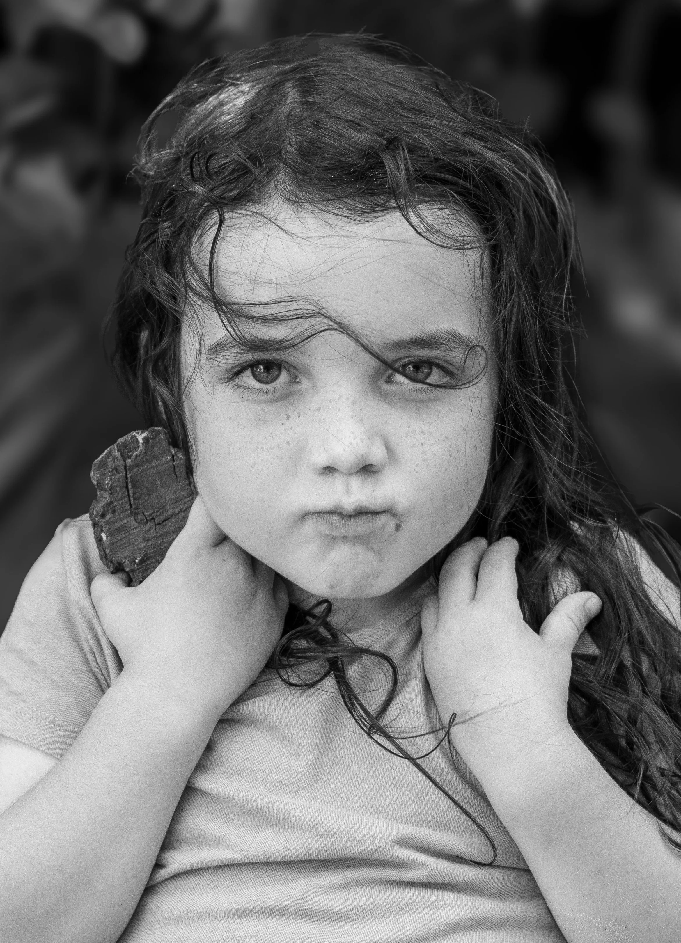 Black and white photo of a young girl with curly hair holding a piece of wood near her neck, looking directly at the camera with a serious expression.
