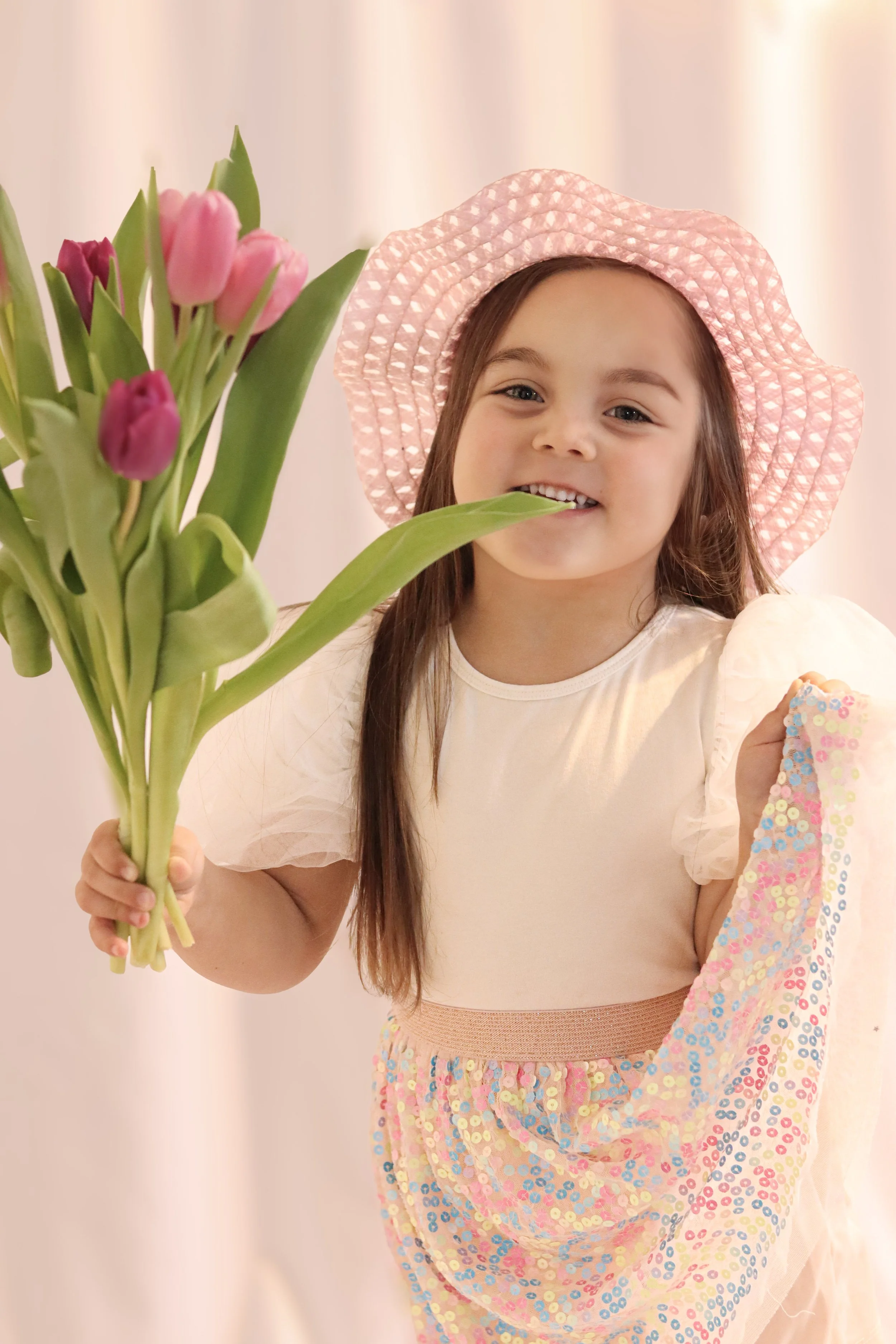 A young girl wearing a pink sunhat and a soft pastel dress holding a bouquet of pink and purple tulips, smiling at the camera.