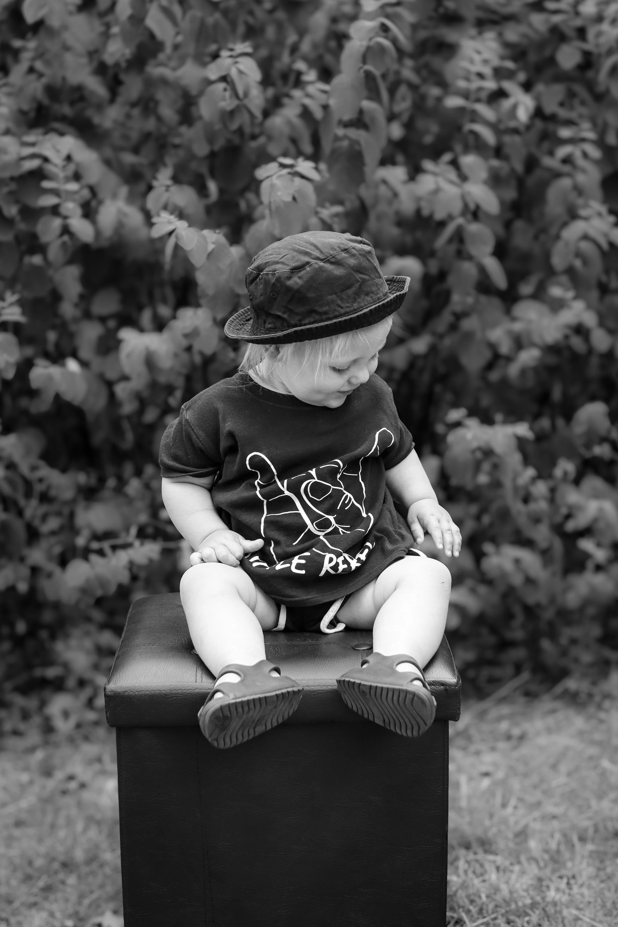 A young child with light-colored hair wearing a black bucket hat, a black t-shirt with a line drawing of a face, shorts, and sandals, sitting on a black box outdoors in front of leafy bushes.