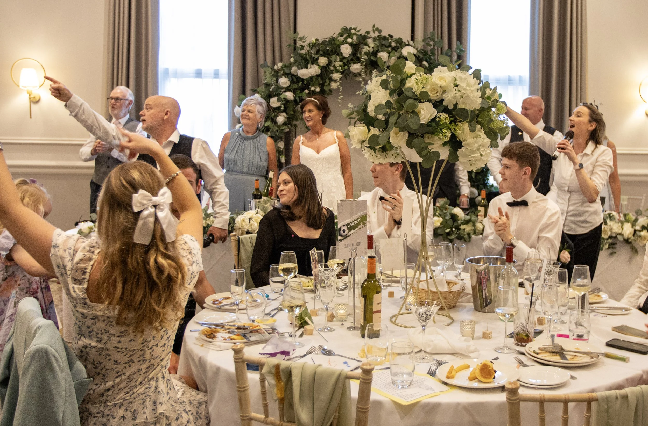 A wedding reception with guests seated at a decorated table and a band performing in the background. The bride is in a white dress, smiling, amidst family and friends.