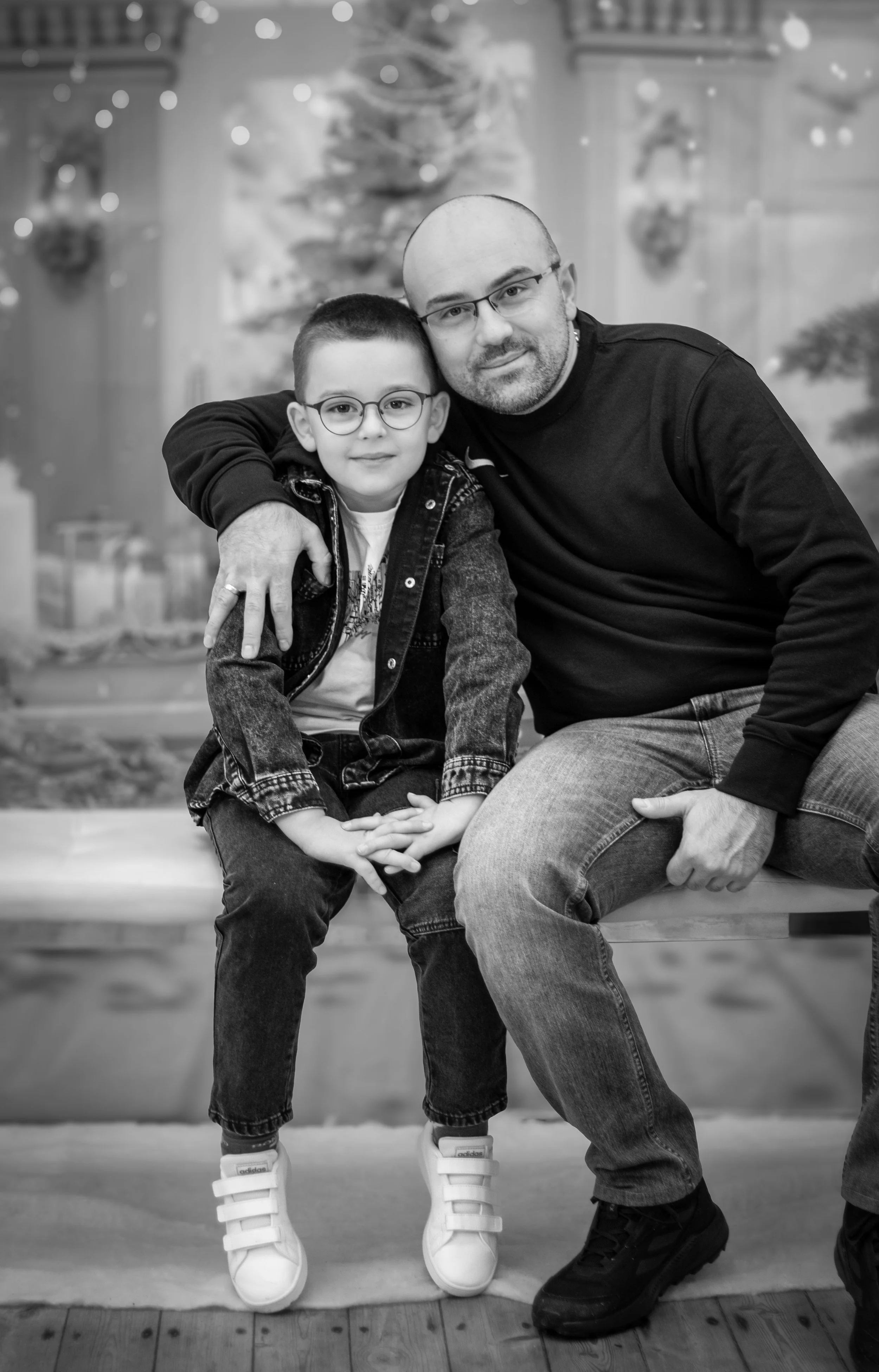 A man and a young boy sitting together on a bench, smiling at the camera, with a Christmas tree in the background.