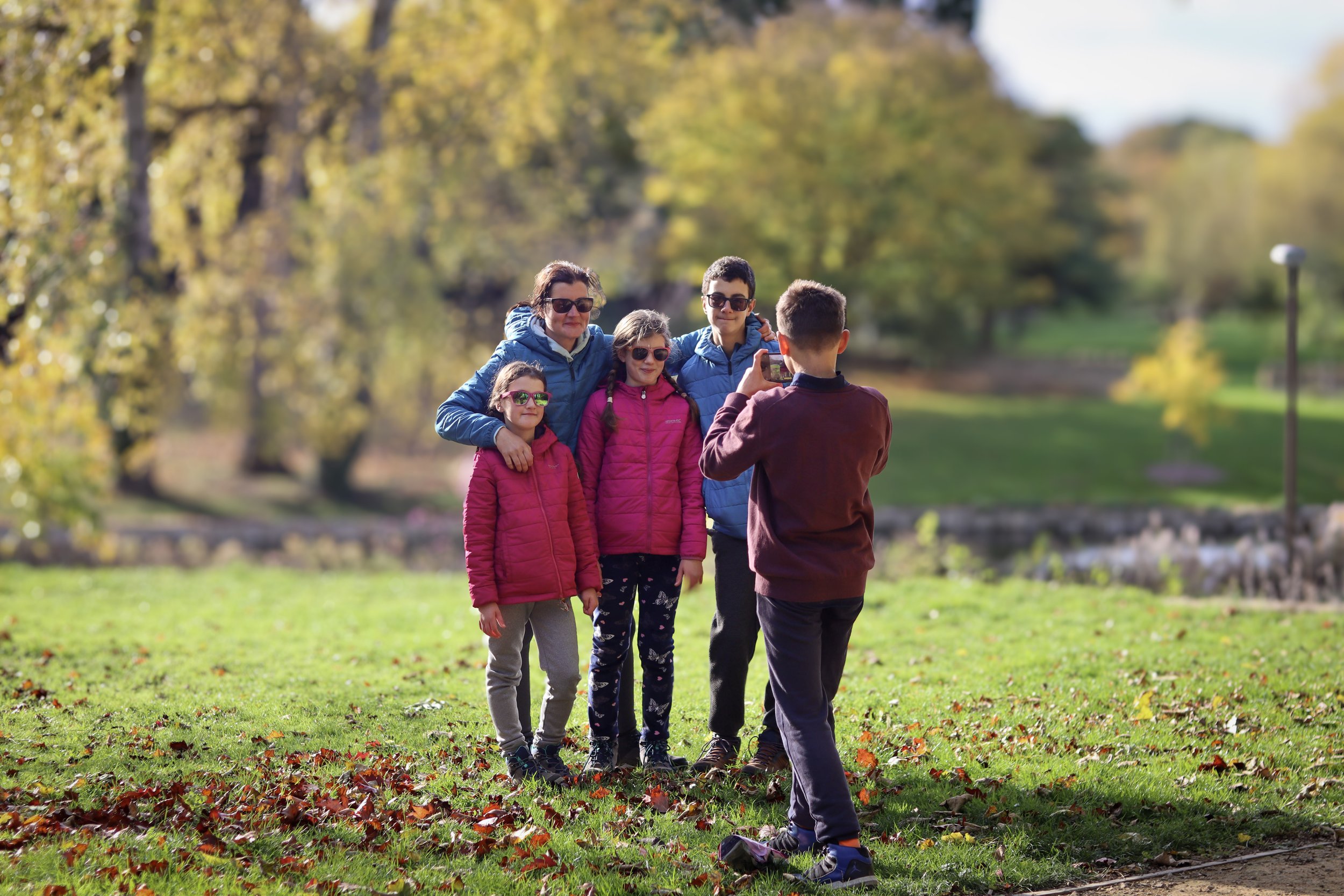 A family of five posing for a photo in a park during autumn, with trees in the background and fallen leaves on the grass.