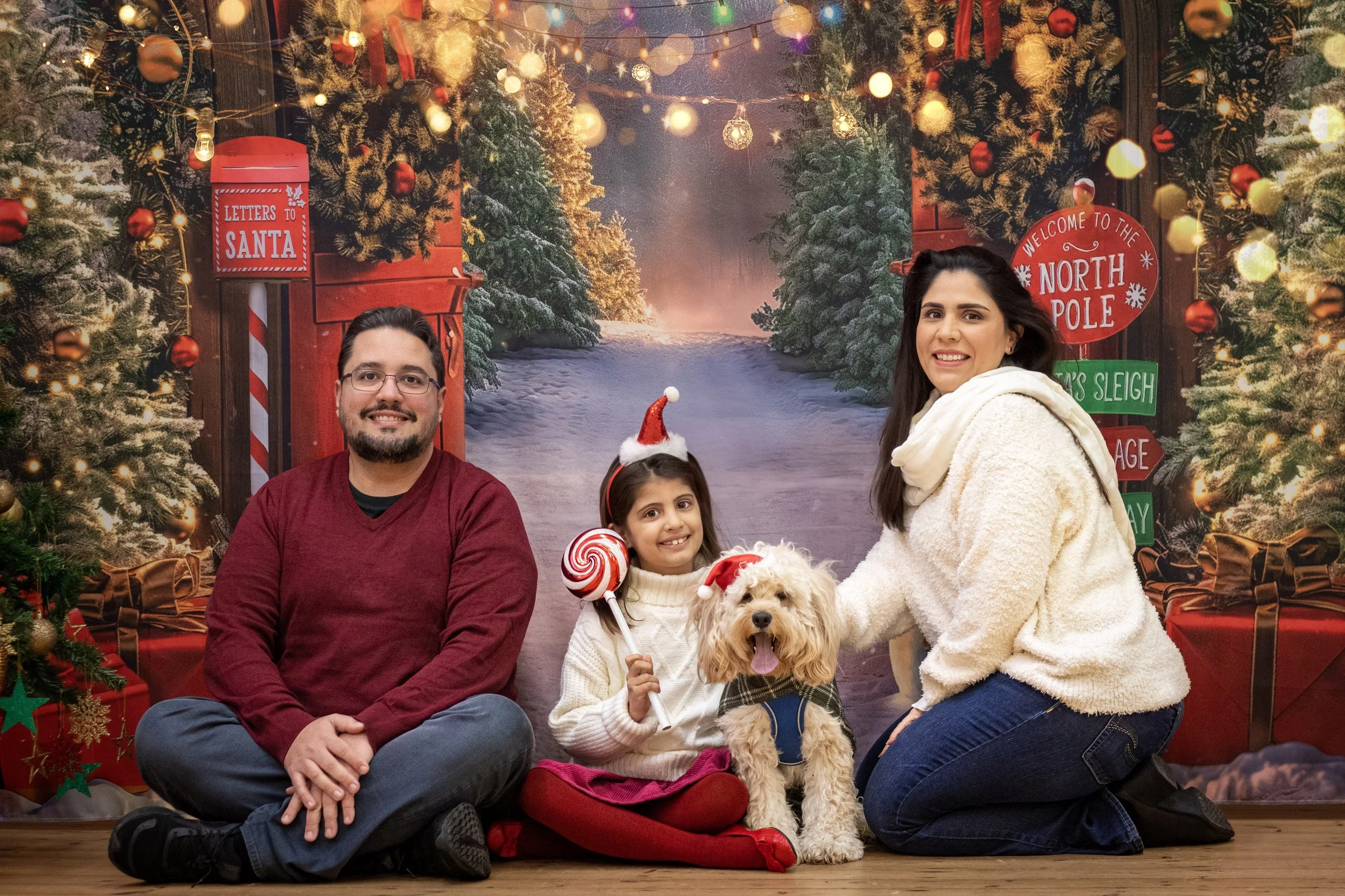 A family of three with a dog posing in front of a festive Christmas backdrop. The backdrop features decorated Christmas trees, pine garlands, hanging lights, and holiday signs. The girl is wearing a Santa hat, holding a lollipop, with the dog wearing a Santa hat beside her. The man and woman are kneeling on the floor smiling at the camera.