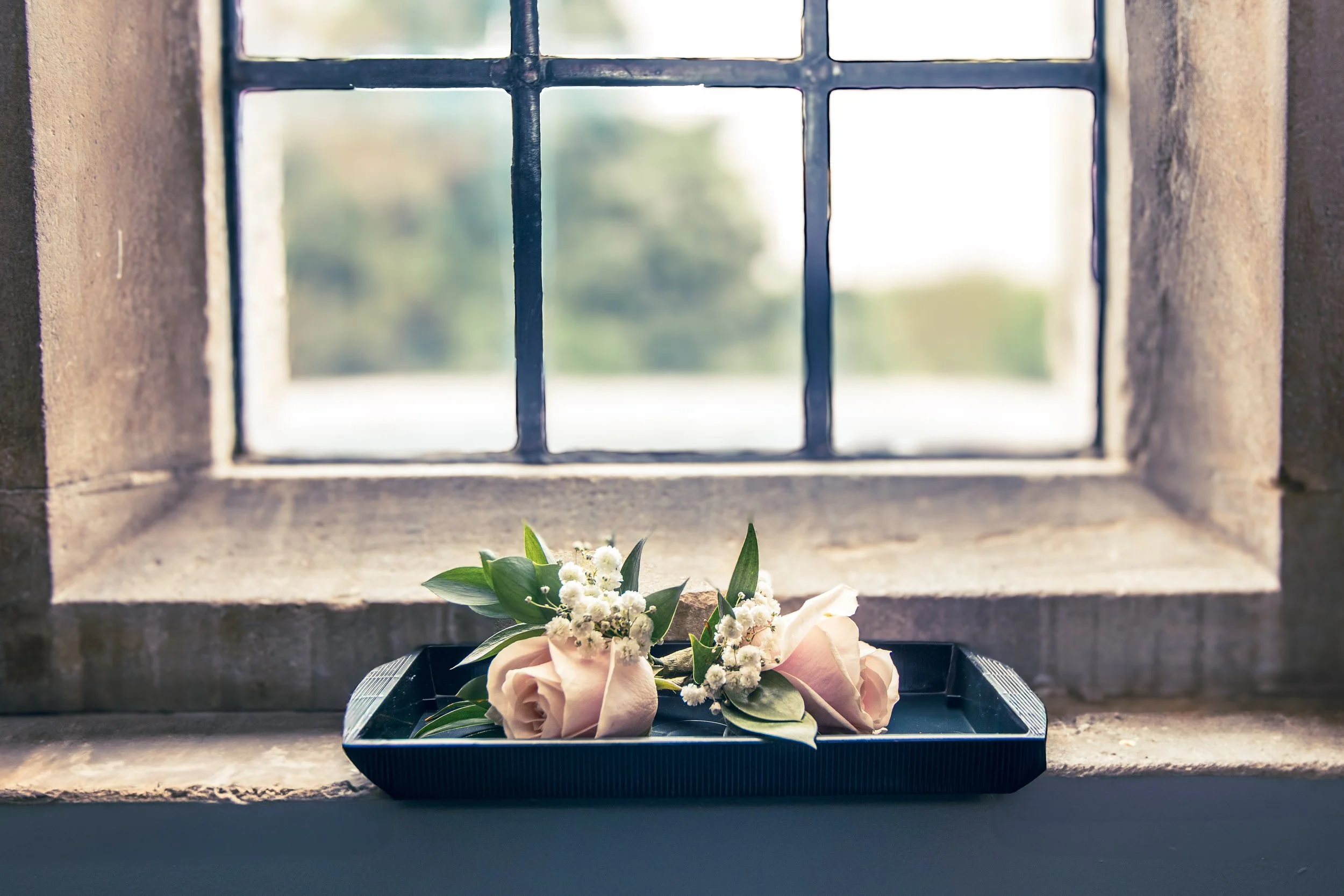 Two pale pink roses with white baby's breath and green leaves on a black tray, placed on a stone window sill, with a large window featuring metal grid in the background showing blurred outdoor scenery.