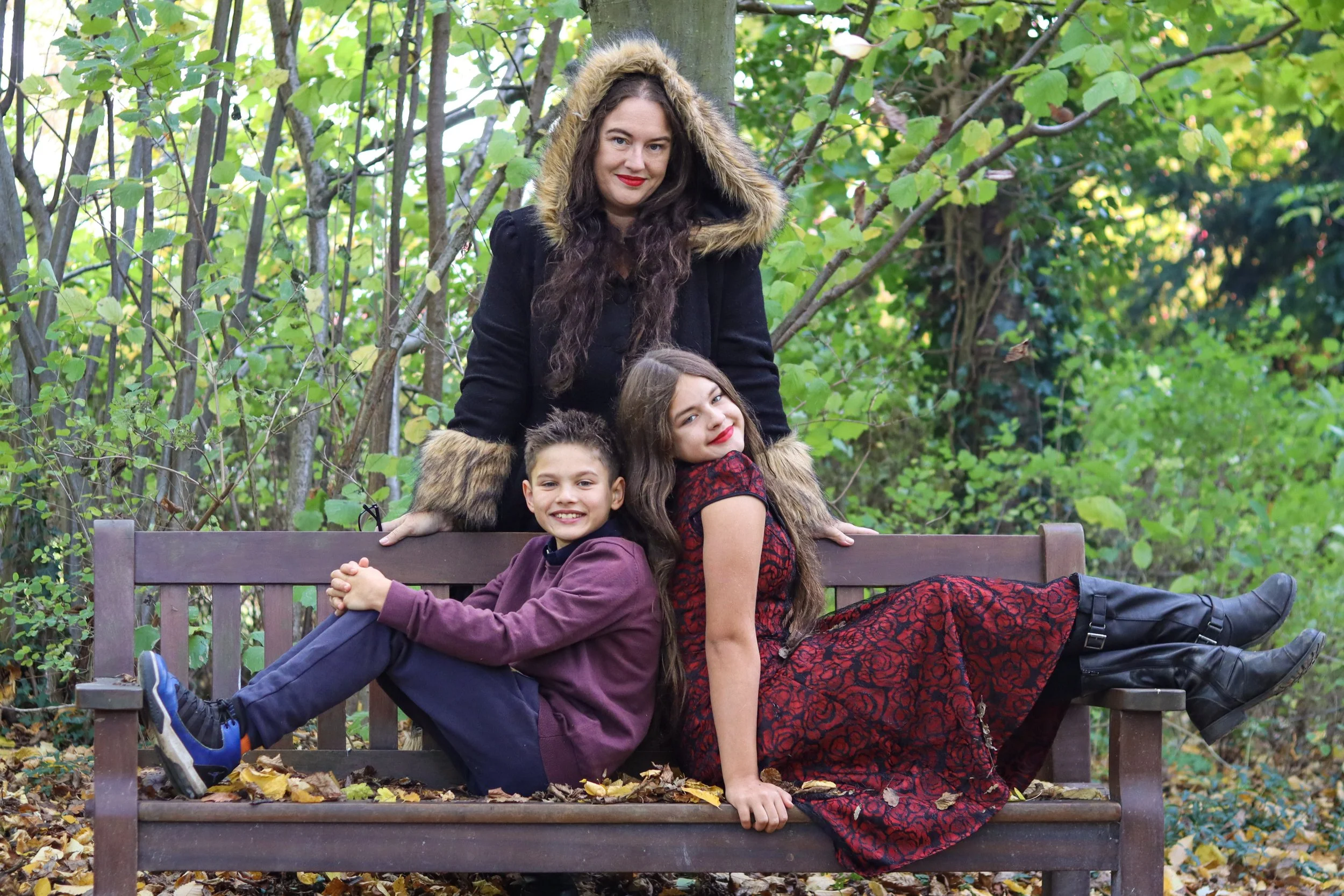 A woman and two children on a park bench surrounded by green trees and fallen leaves. The woman is standing behind the bench, wearing a black coat with a fur-lined hood, and has long wavy hair and red lipstick. The girl is sitting on the bench, leaning against the woman, with long brown hair, a red and black dress, and boots. The boy is sitting on the left side of the bench, wearing a purple jacket, dark pants, and sneakers. All are smiling and enjoying the outdoor setting.