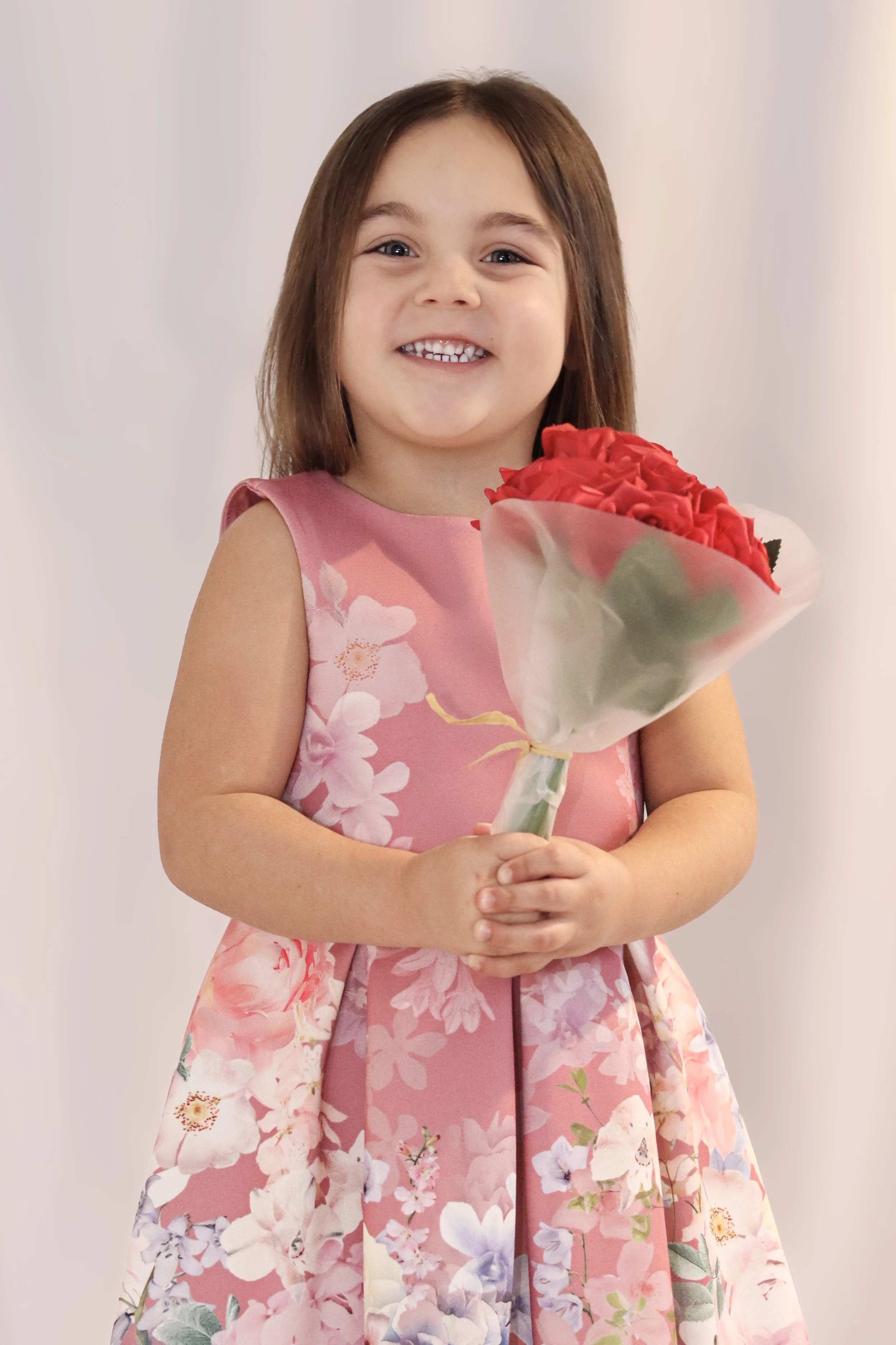 A young girl with brown hair smiling and holding a bouquet of red roses, wearing a pink floral dress.