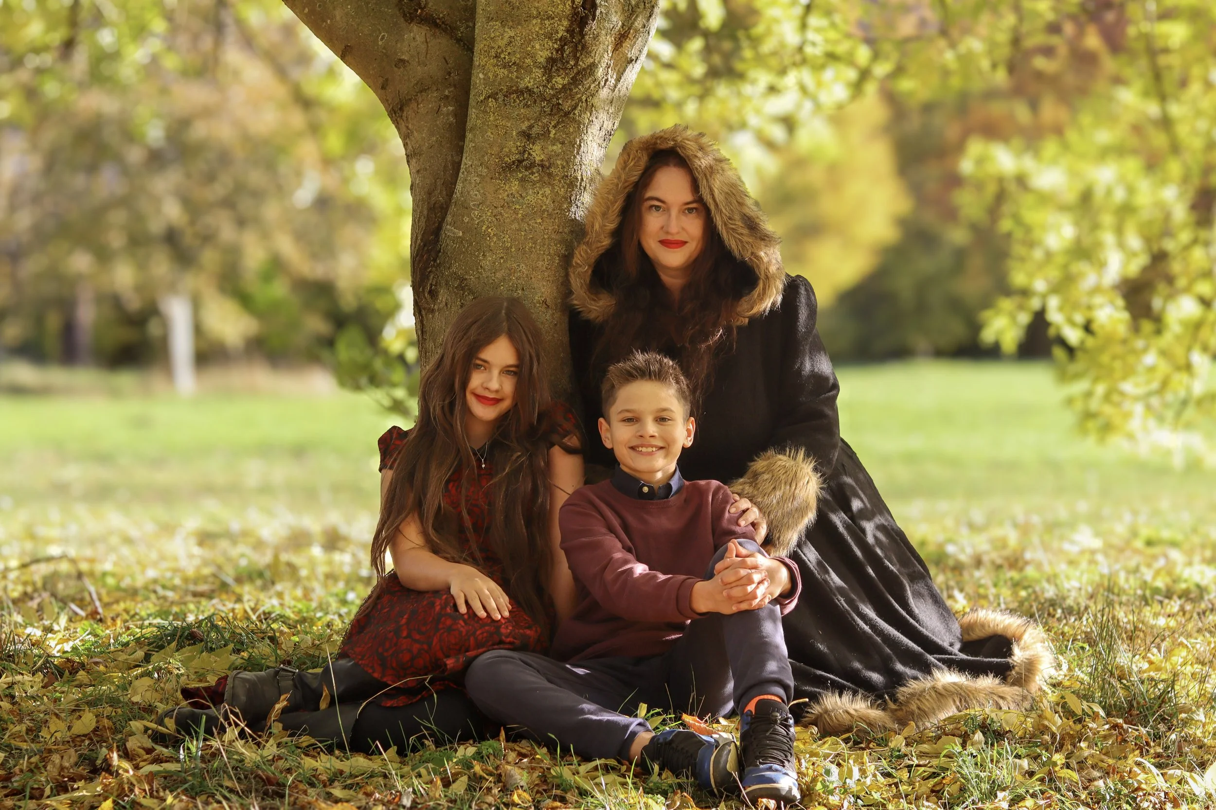 A woman with long, dark hair and a furry hooded coat sitting under a tree with a boy and girl, both children with long hair, in a park during fall, surrounded by yellow and green leaves.