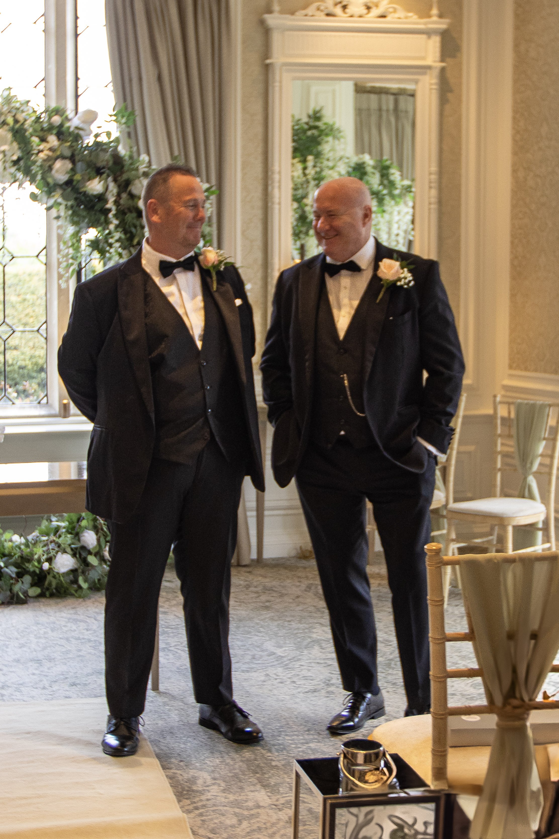 Two men in tuxedos with boutonnieres smiling and talking inside a decorated room, possibly at a wedding or formal event.