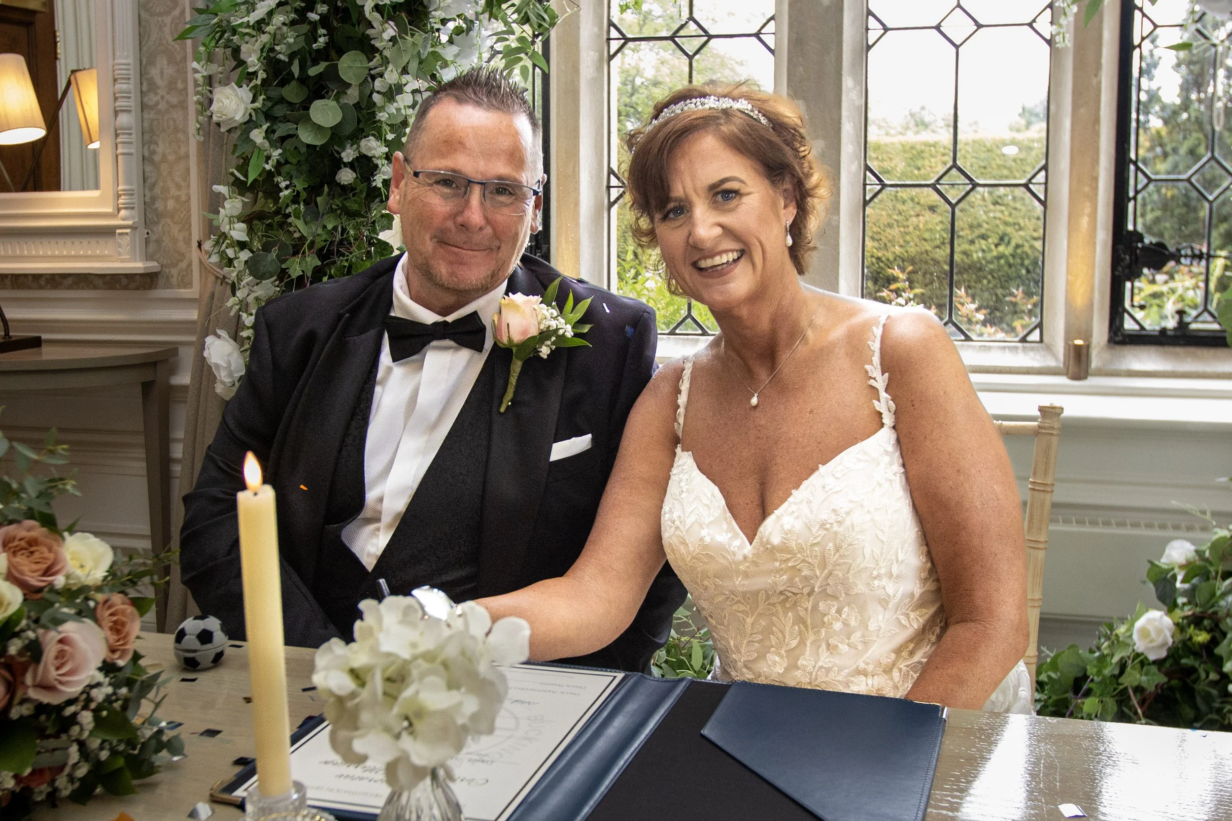 A bride and groom sitting at a wedding reception table, smiling at the camera, with wedding decorations and a window in the background.