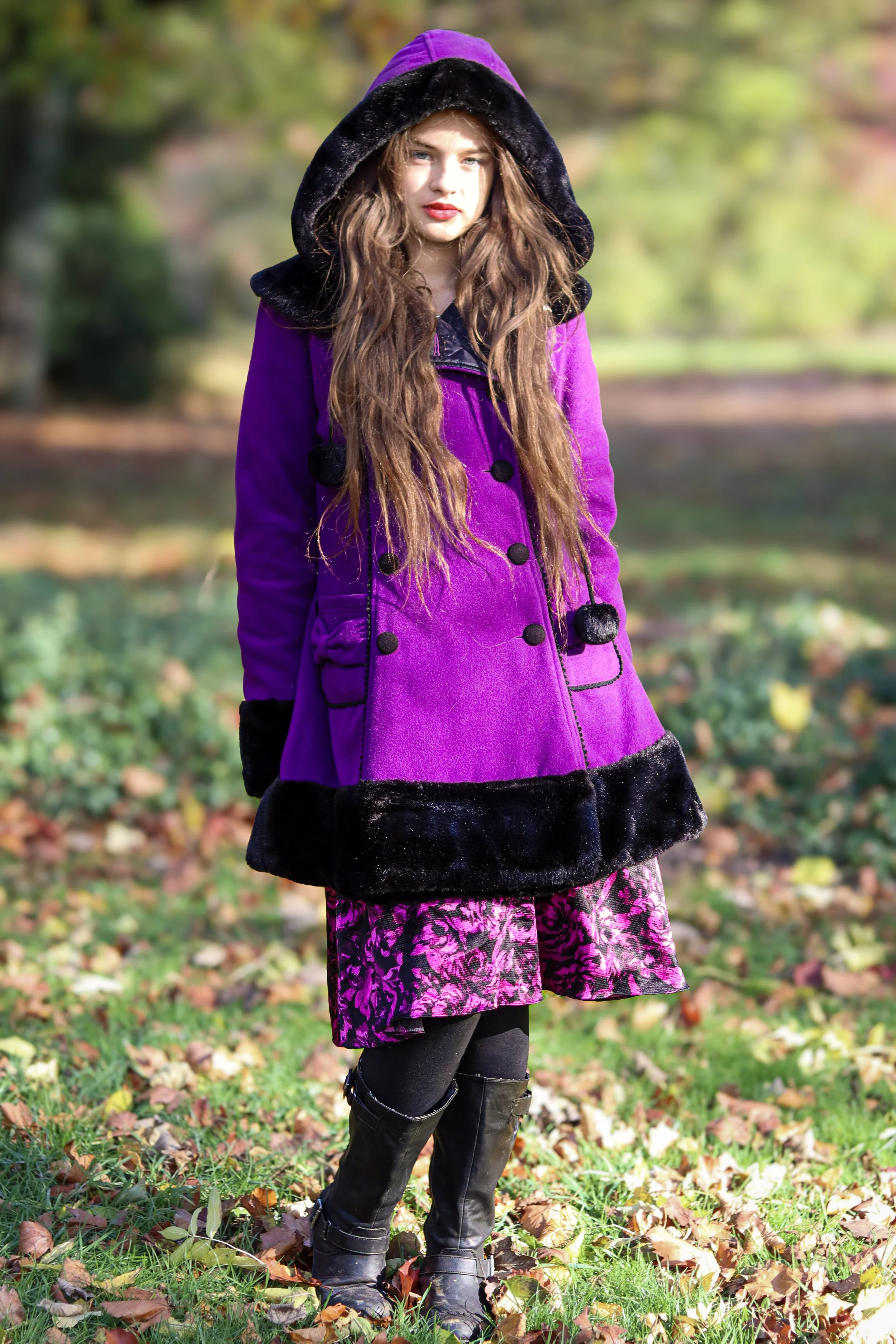 A woman with long brown hair stands outdoors among fallen autumn leaves, wearing a purple coat with black fur trim and a purple and black patterned skirt, with black tights and tall black boots. The background features blurred trees and greenery.