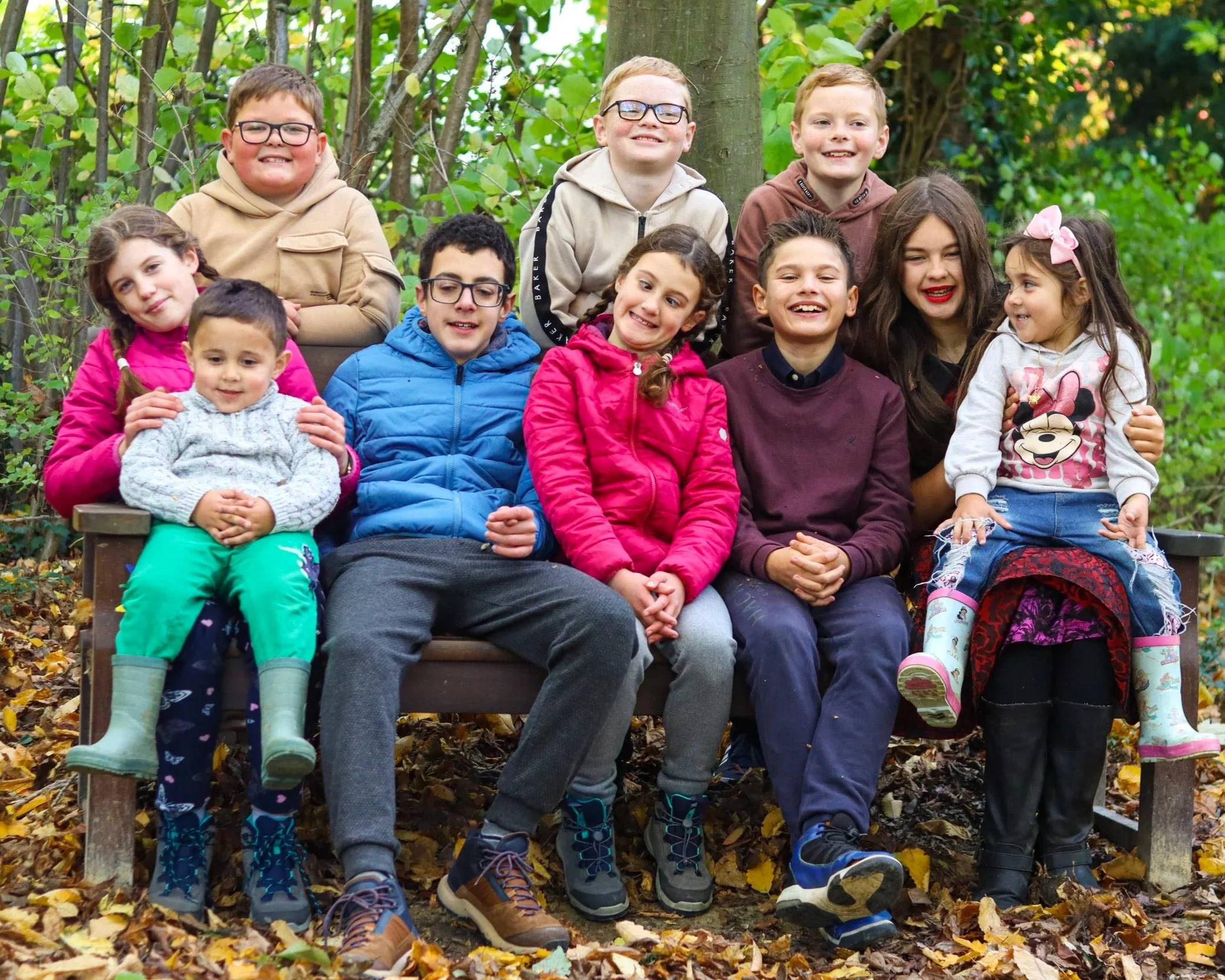 Group of children sitting and standing on a park bench surrounded by fallen leaves and green trees, smiling and enjoying a sunny day.