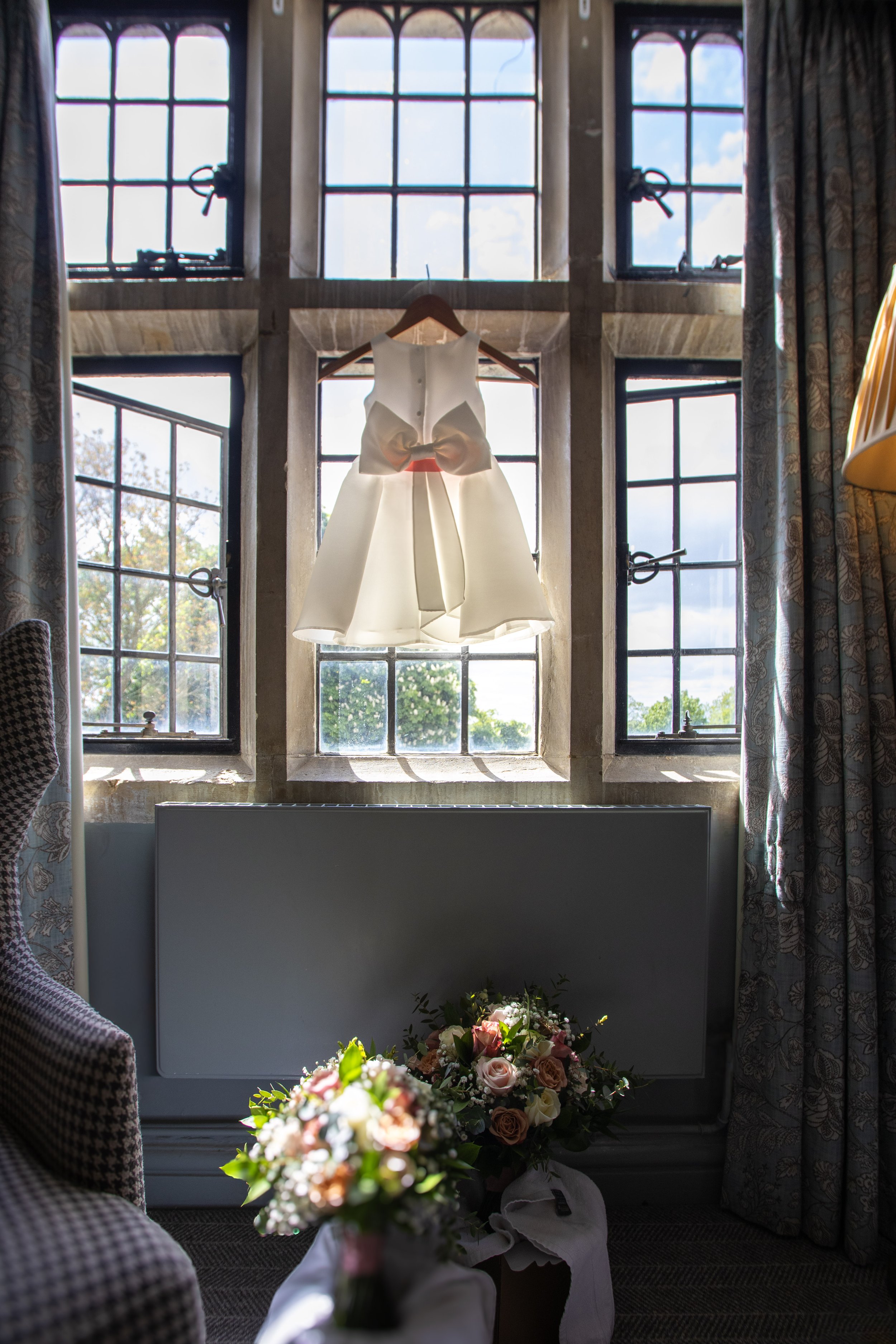 A white dress with a bow detail hanging in front of a window with sunlight streaming through, with bouquets of flowers on the floor below.