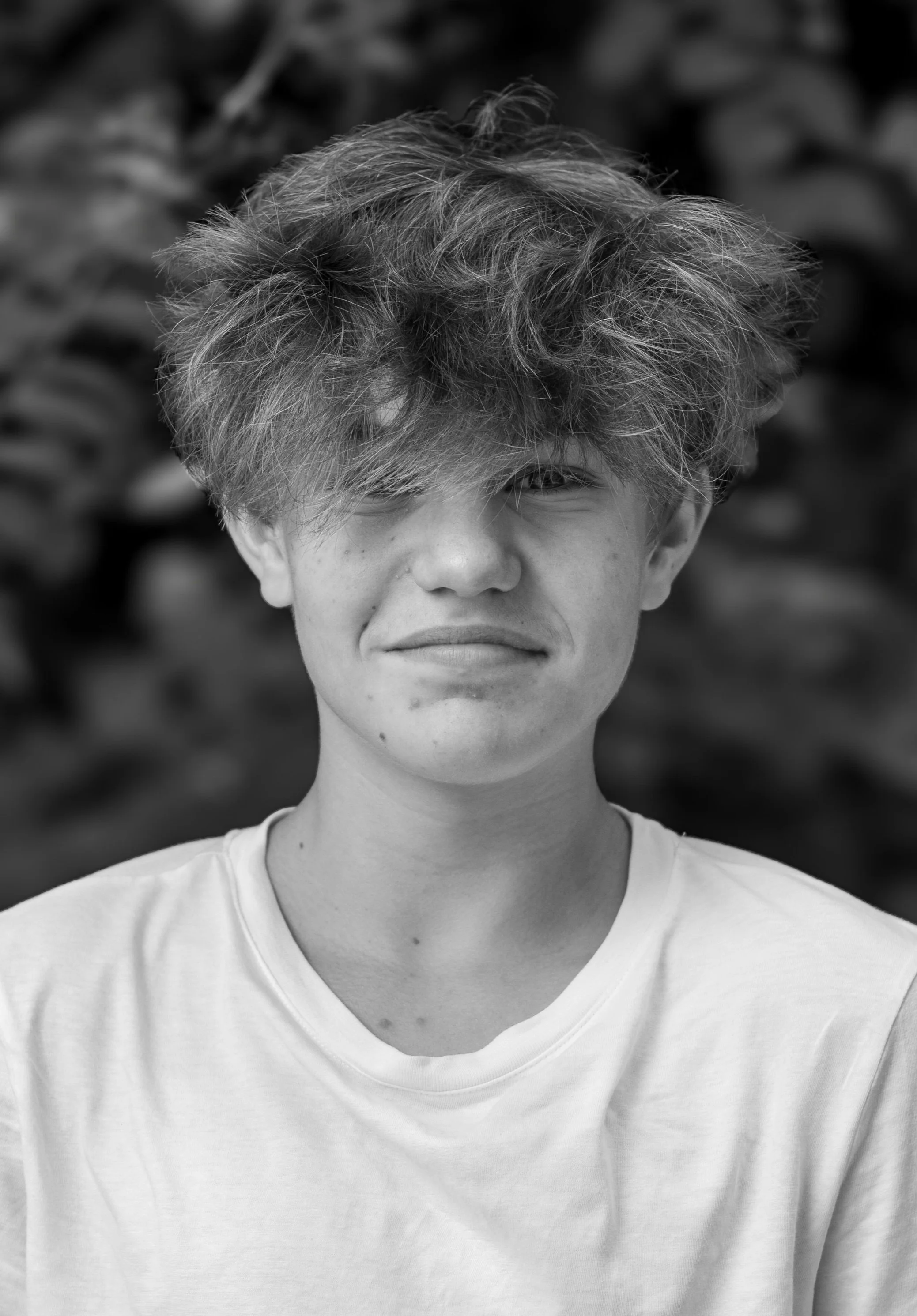 Black and white photo of a young man with curly, tousled hair, wearing a white T-shirt, standing outdoors with leafy background.