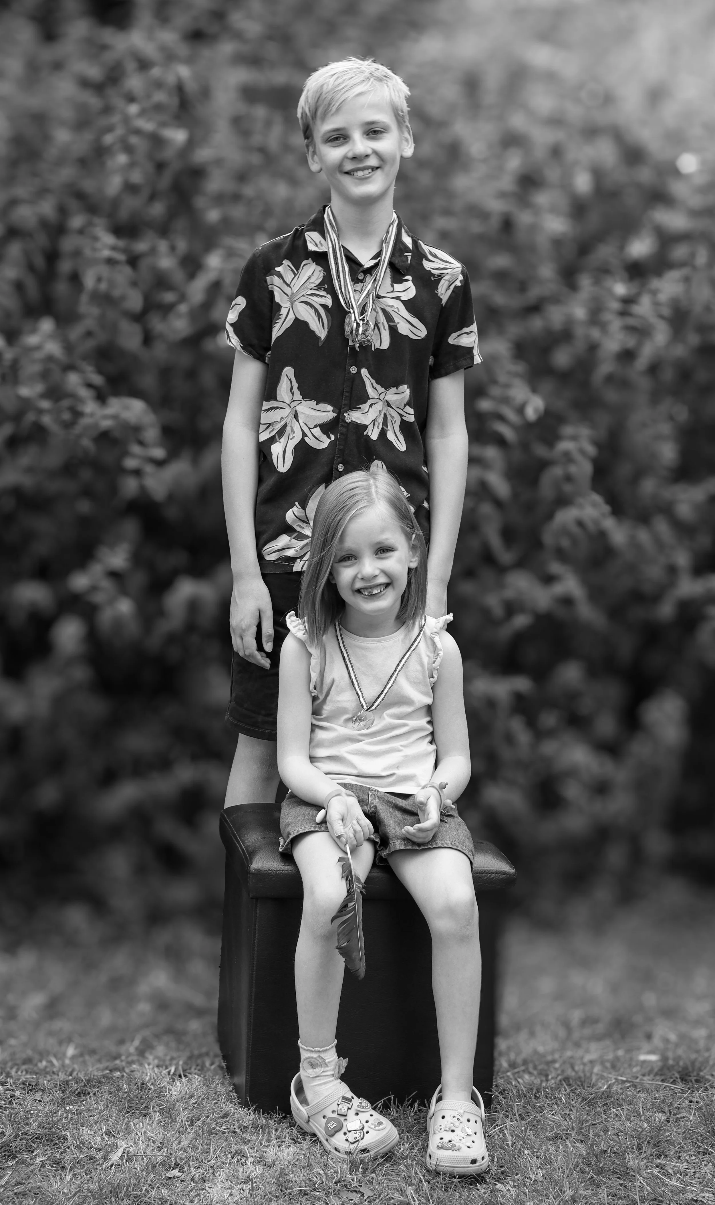 A black and white photo of a boy and a girl, with the boy standing behind a seated girl, both smiling and wearing medals around their necks, outdoors with blurred foliage in the background.