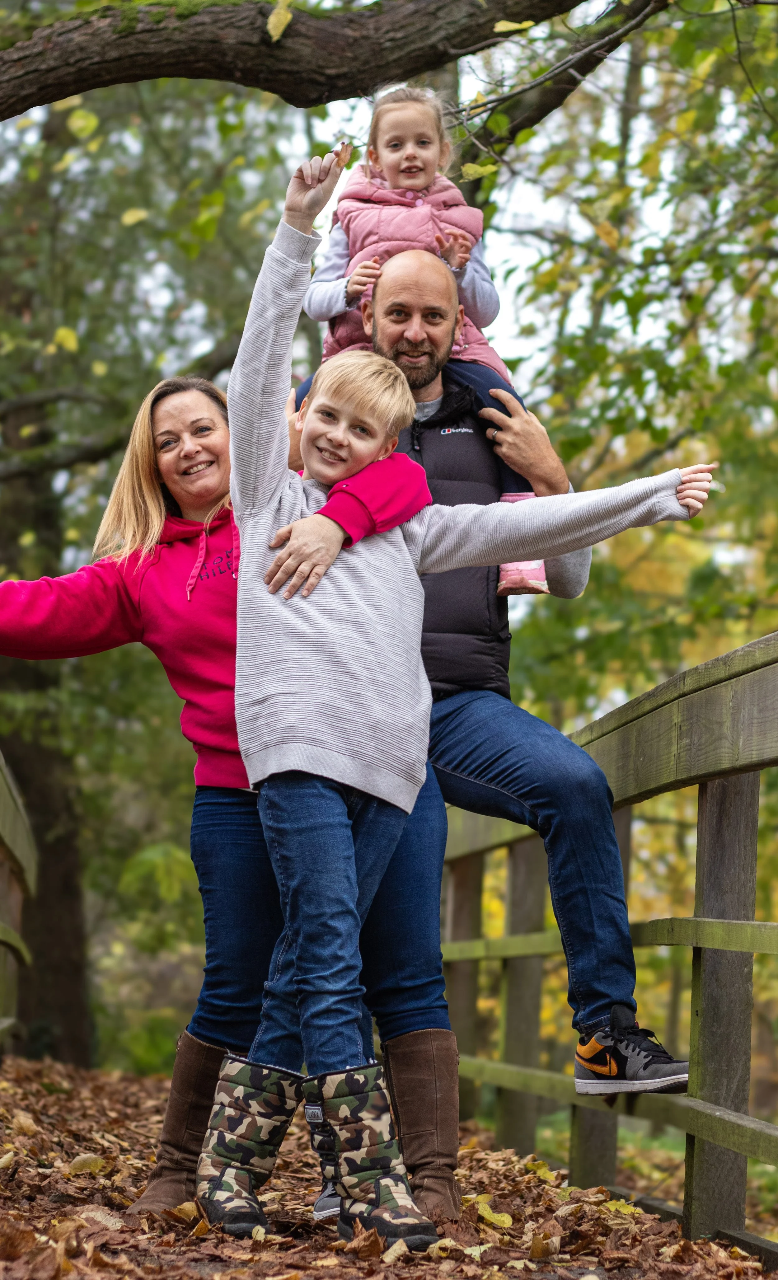 A family of four outdoors in a park during fall, with the father carrying a young girl on his shoulders, the mother standing next to him, and an older boy standing in front with arms outstretched.
