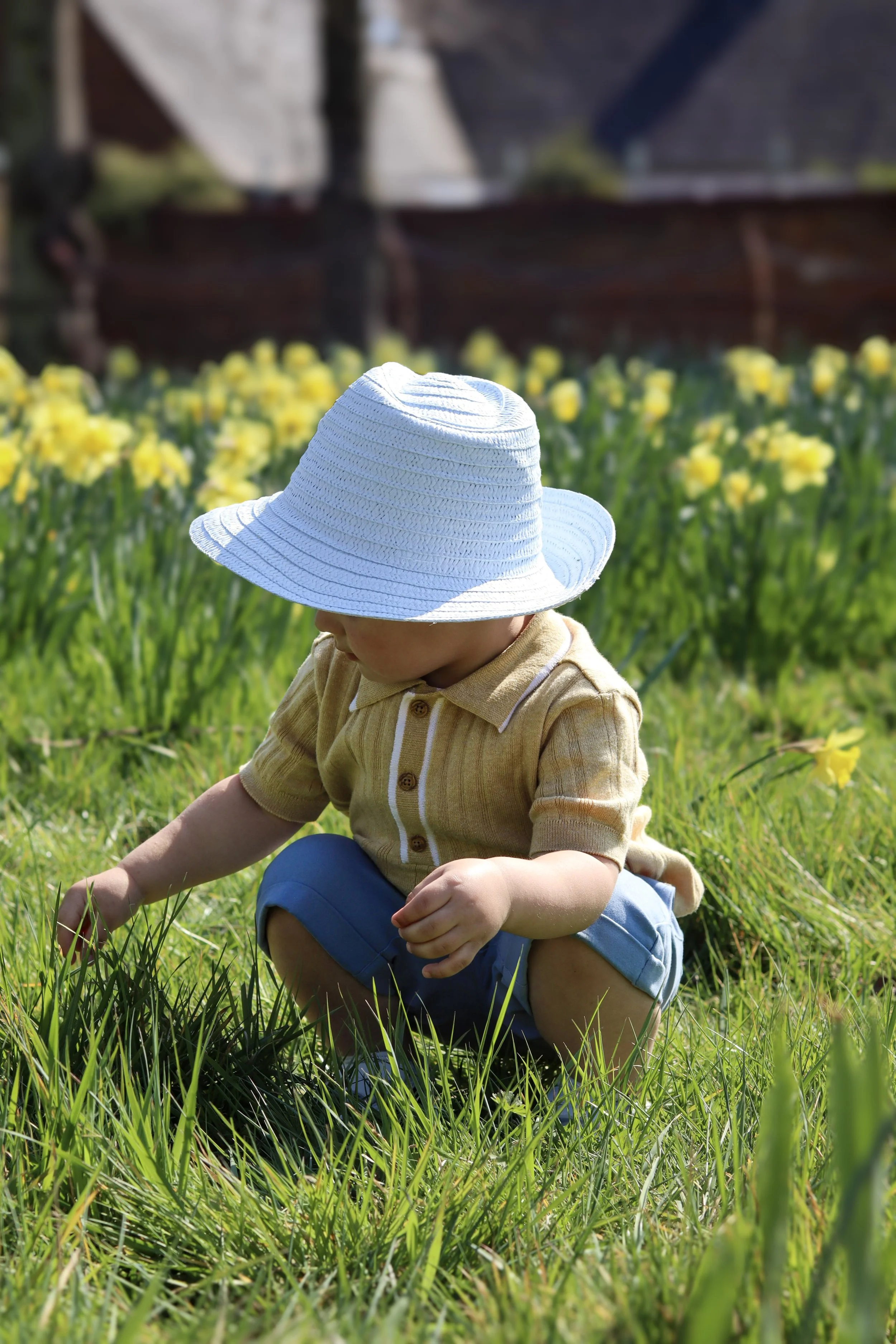 Child wearing a white sun hat, tan shirt, and blue shorts squatting in a grassy field near yellow flowers.