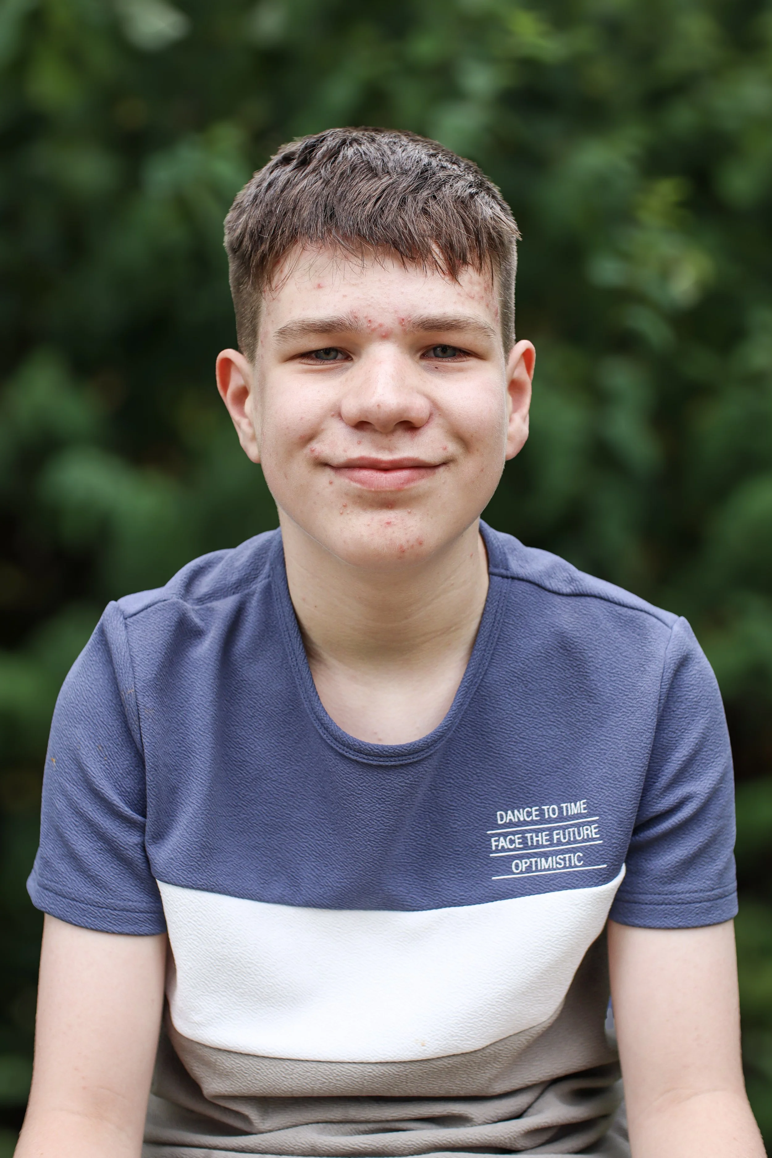 A young boy with short brown hair and acne, wearing a navy blue T-shirt with a white and beige stripe, smiling outdoors with blurred green foliage in the background.