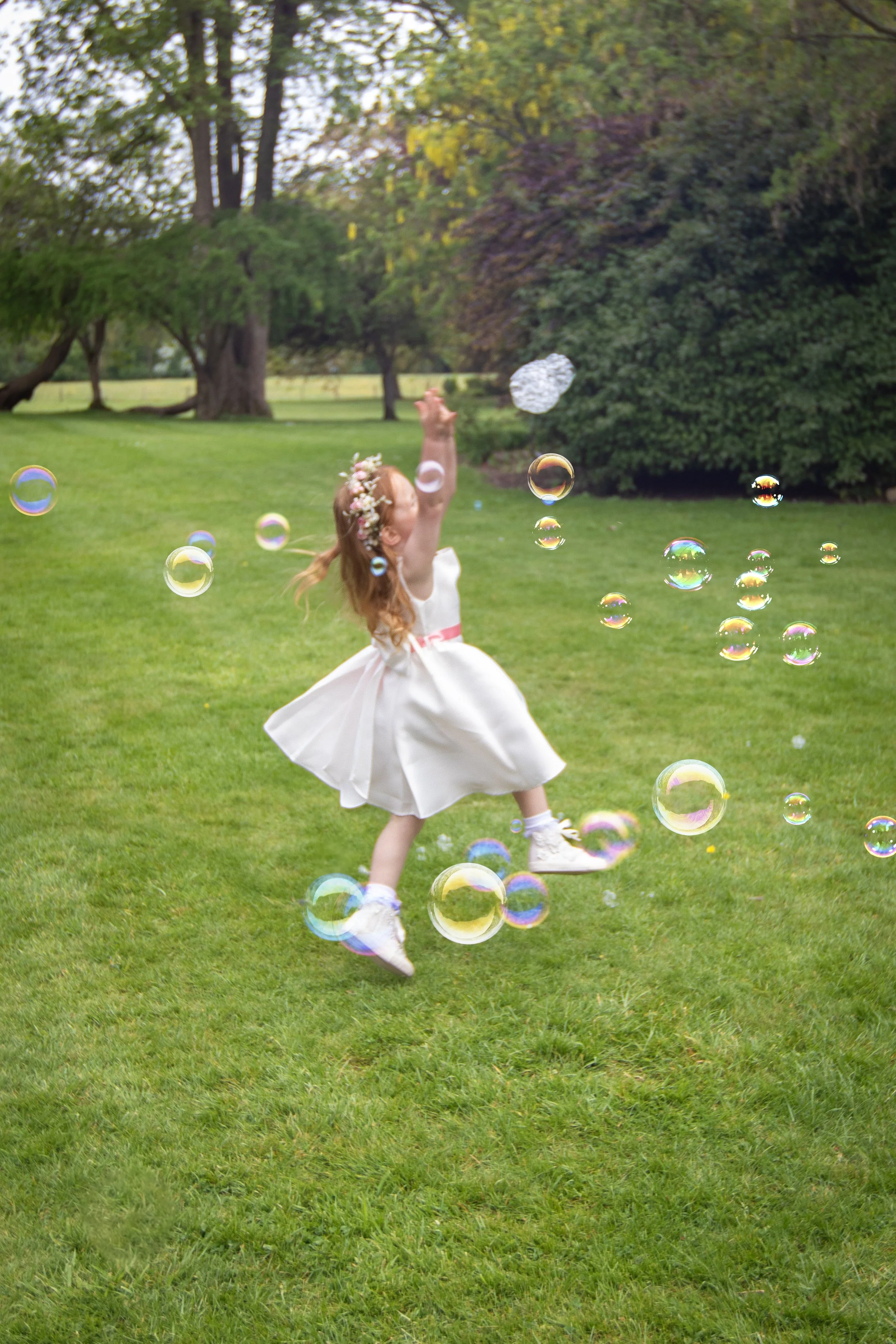 A young girl in a white dress and floral headband playing with soap bubbles outdoors on a grassy field with trees in the background.