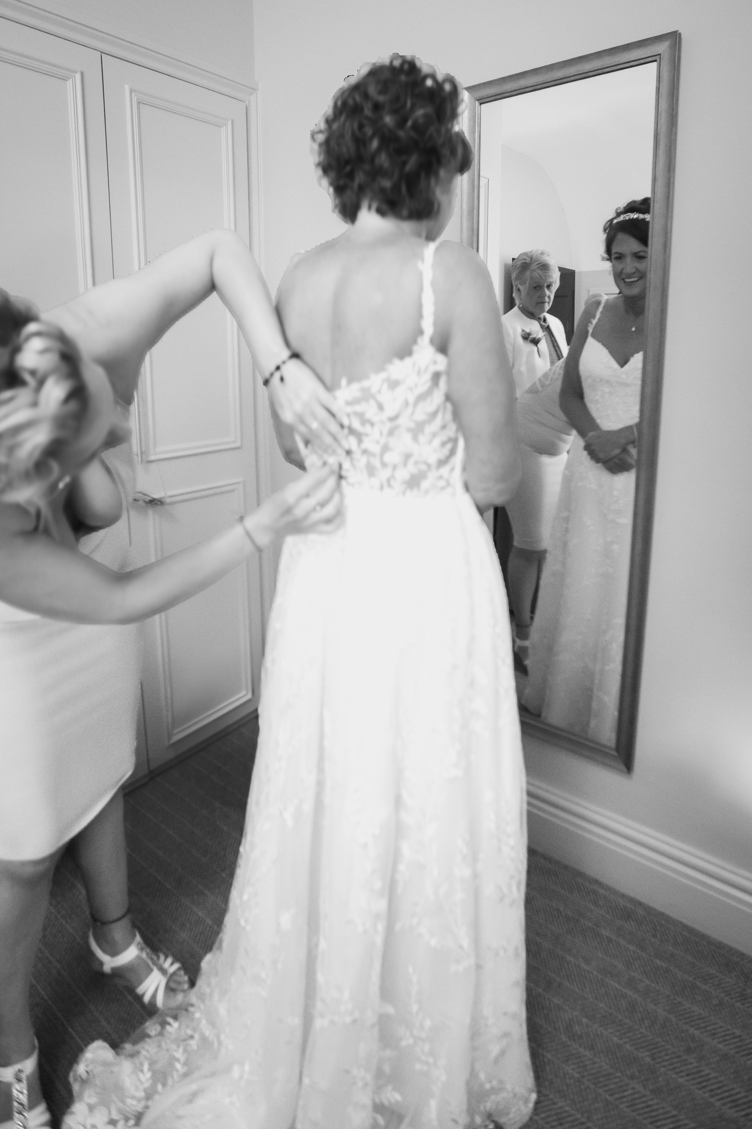 A bride trying on her wedding dress while a helper assists in buttoning the back, with a guest looking on through a mirror.