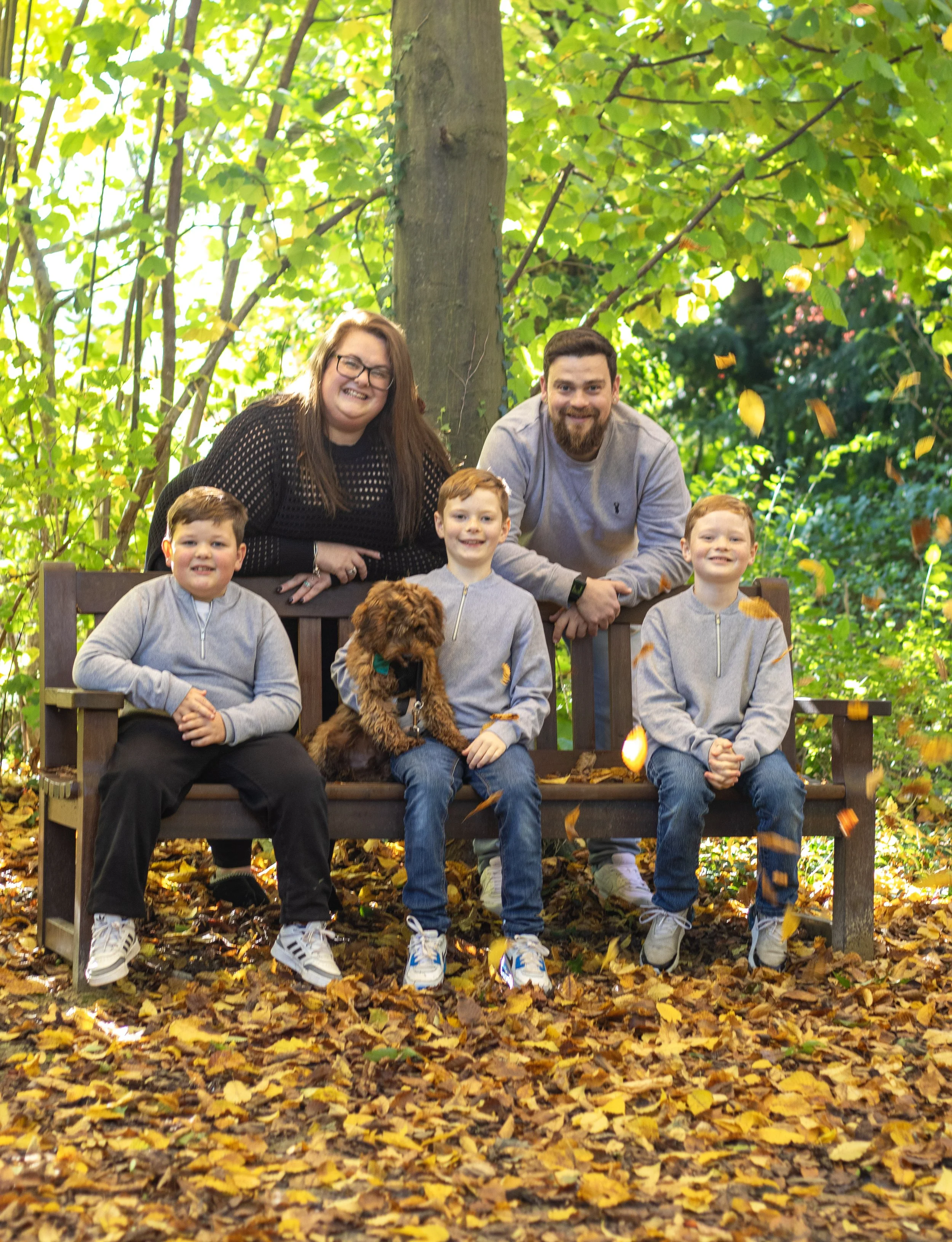 Family of five with four children and a dog sitting on a bench in a wooded park during fall, surrounded by yellow leaves.