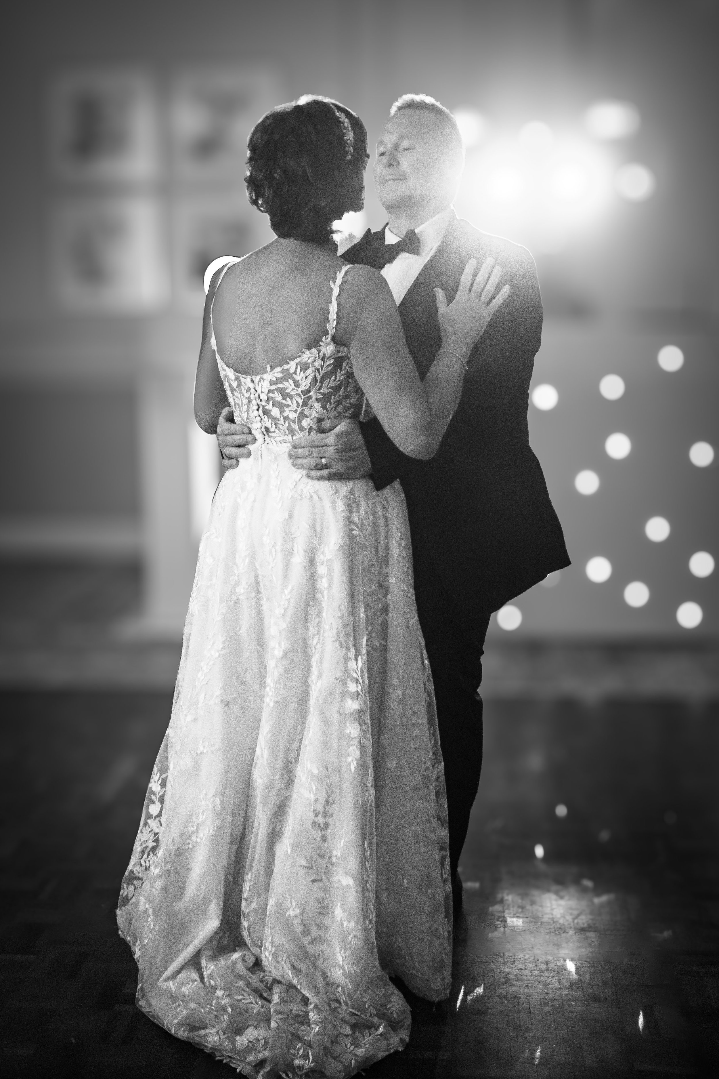 Black and white photo of a wedding dance with a bride in a lace gown and a groom in a tuxedo, dancing closely indoors.