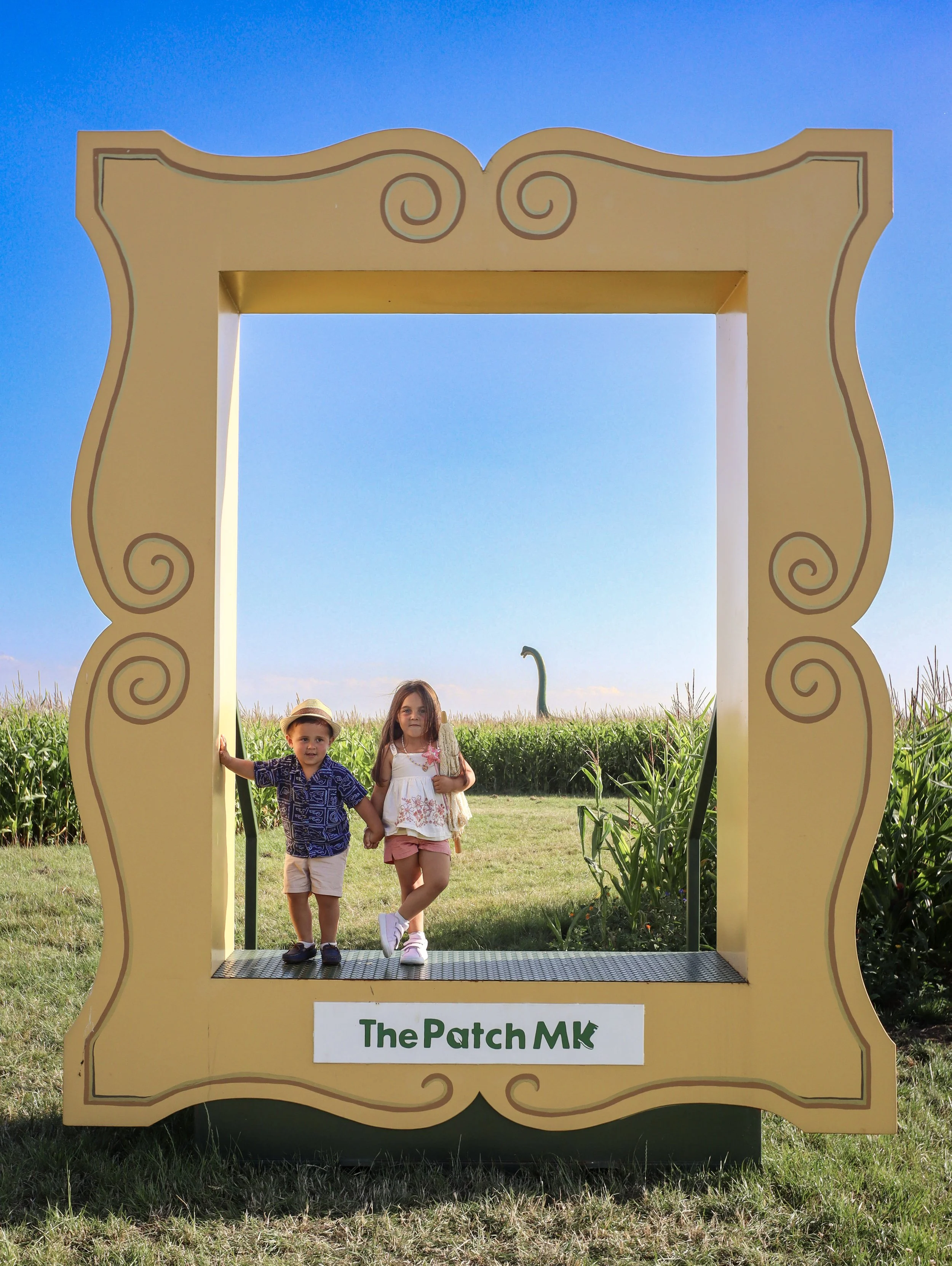 Two children, a boy and a girl, standing in a large decorative frame at The Patch MK, with a field and a dinosaur sculpture in the background, under a clear blue sky.
