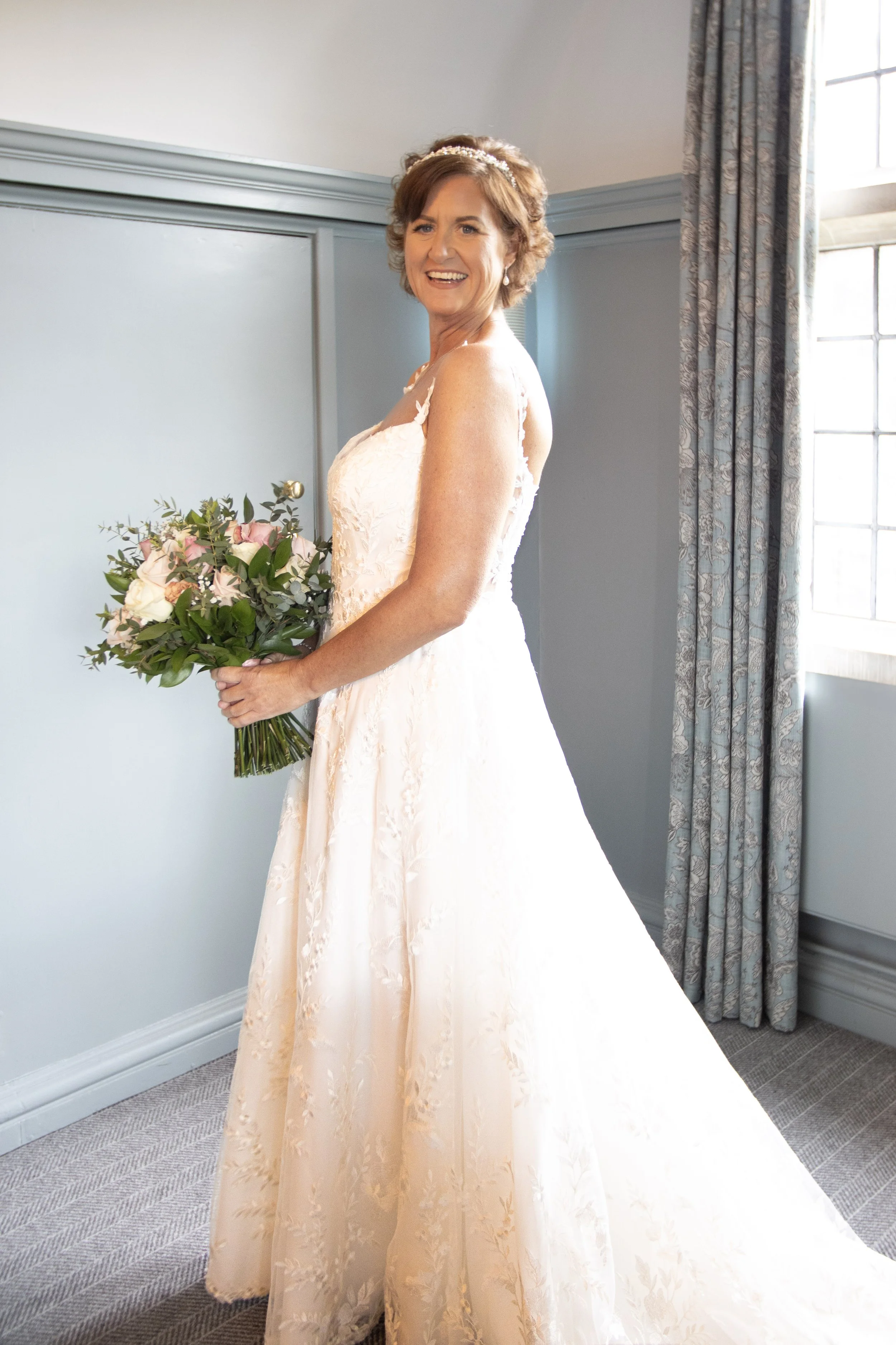 A smiling woman in a white wedding dress holding a bouquet of flowers, standing indoors near a window and curtain.