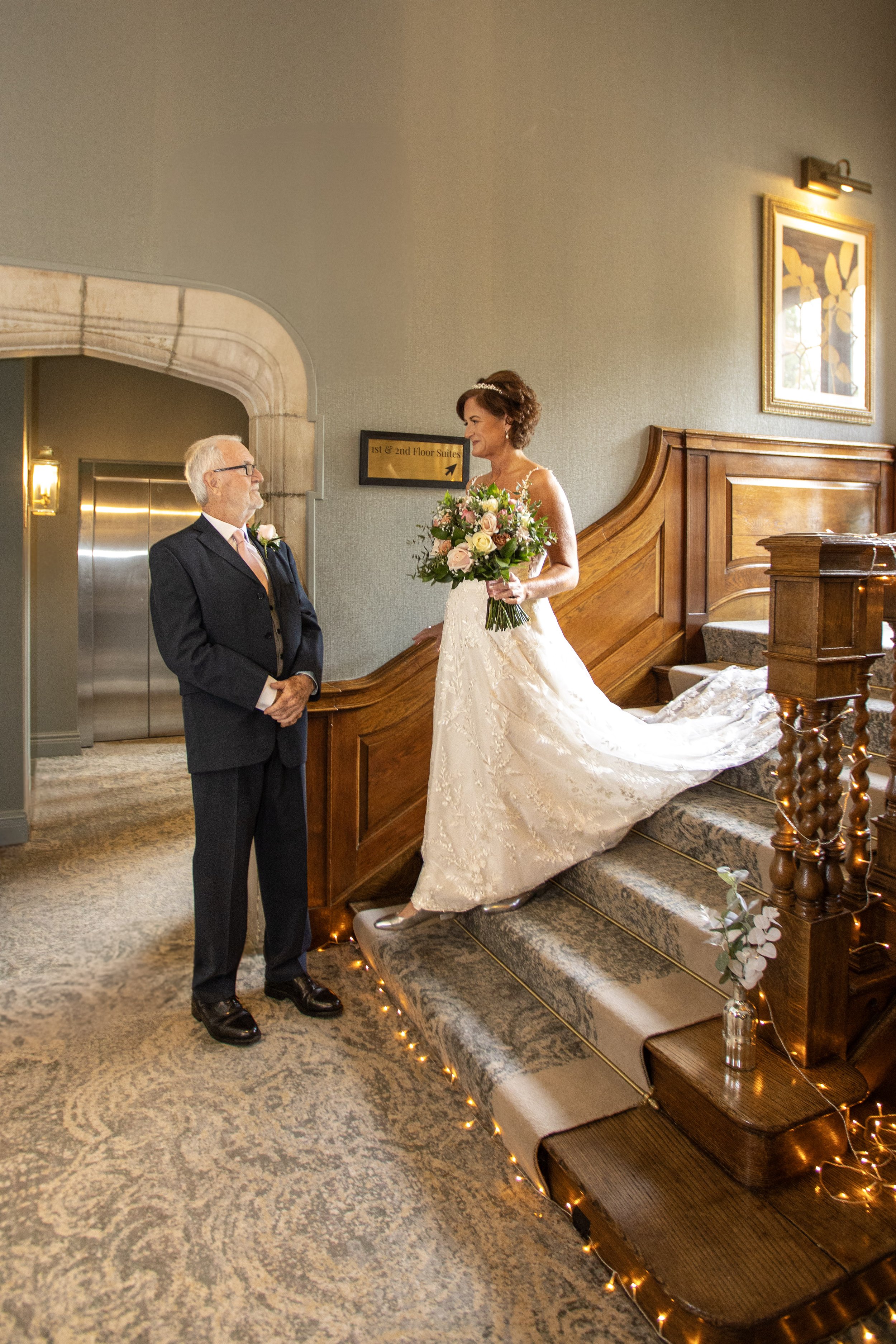 A bride in a white lace wedding gown holding a bouquet of pink roses and greenery, standing on stairs and smiling at a man in a dark suit, inside a decorated room with warm lighting.