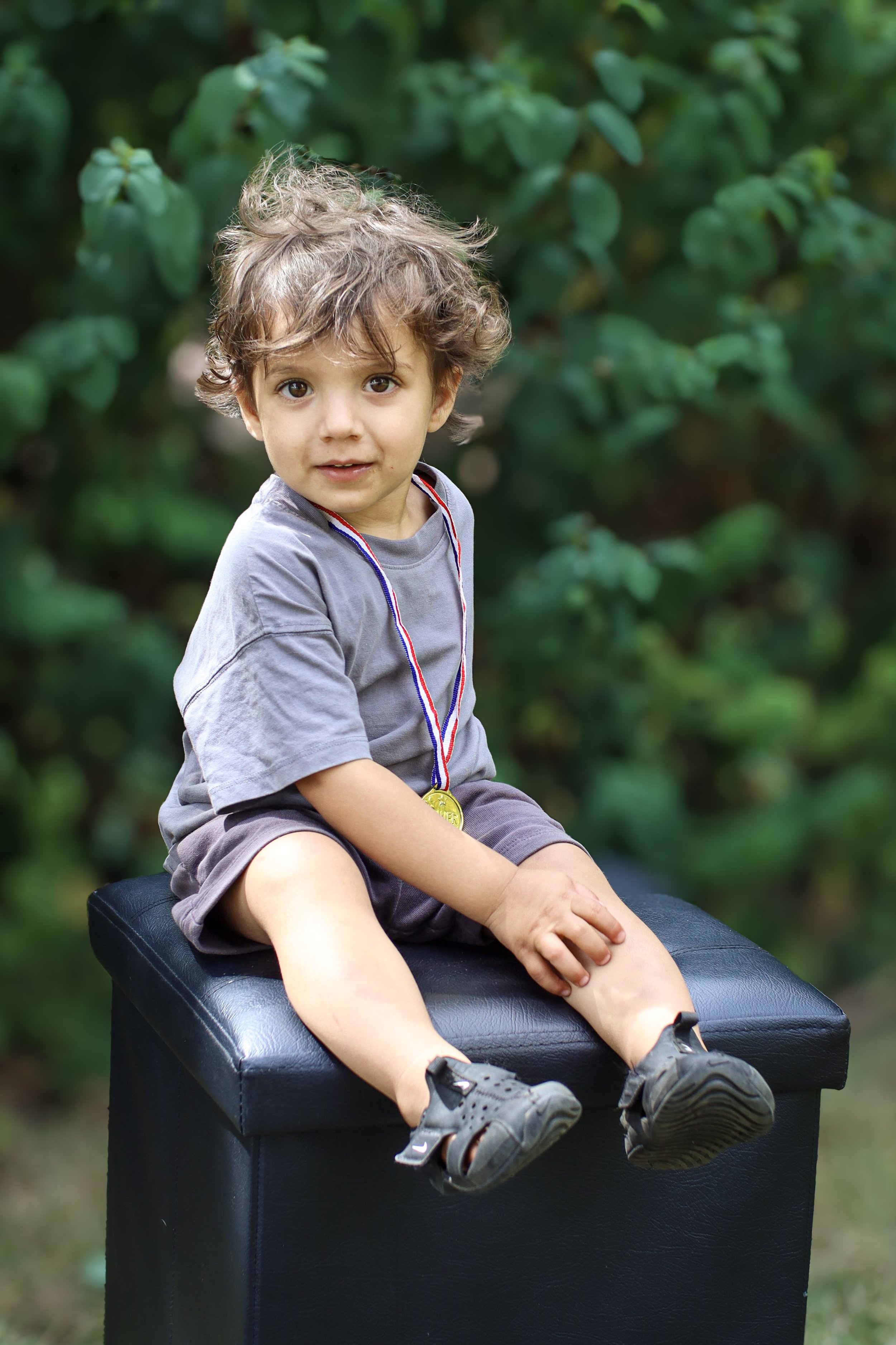 Young boy sitting on a black ottoman outdoors, wearing a gray t-shirt and shorts with a gold medal around his neck, surrounded by green foliage.