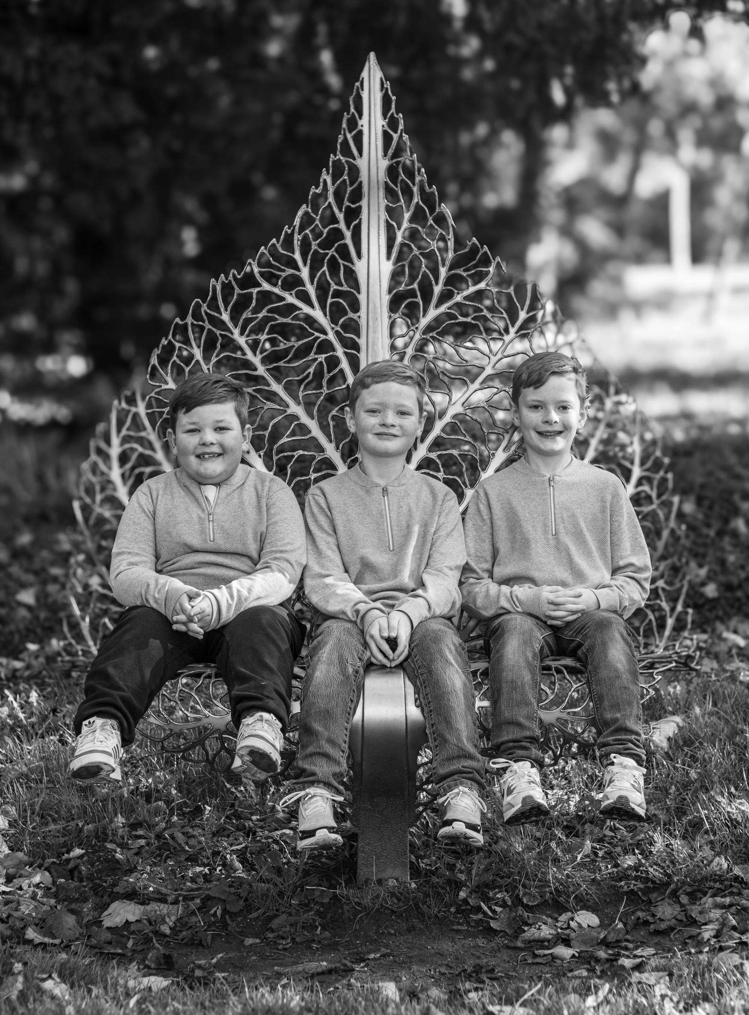 Three young boys sitting on a decorative metal garden bench with large leaf design in the background, outdoors, in black and white.