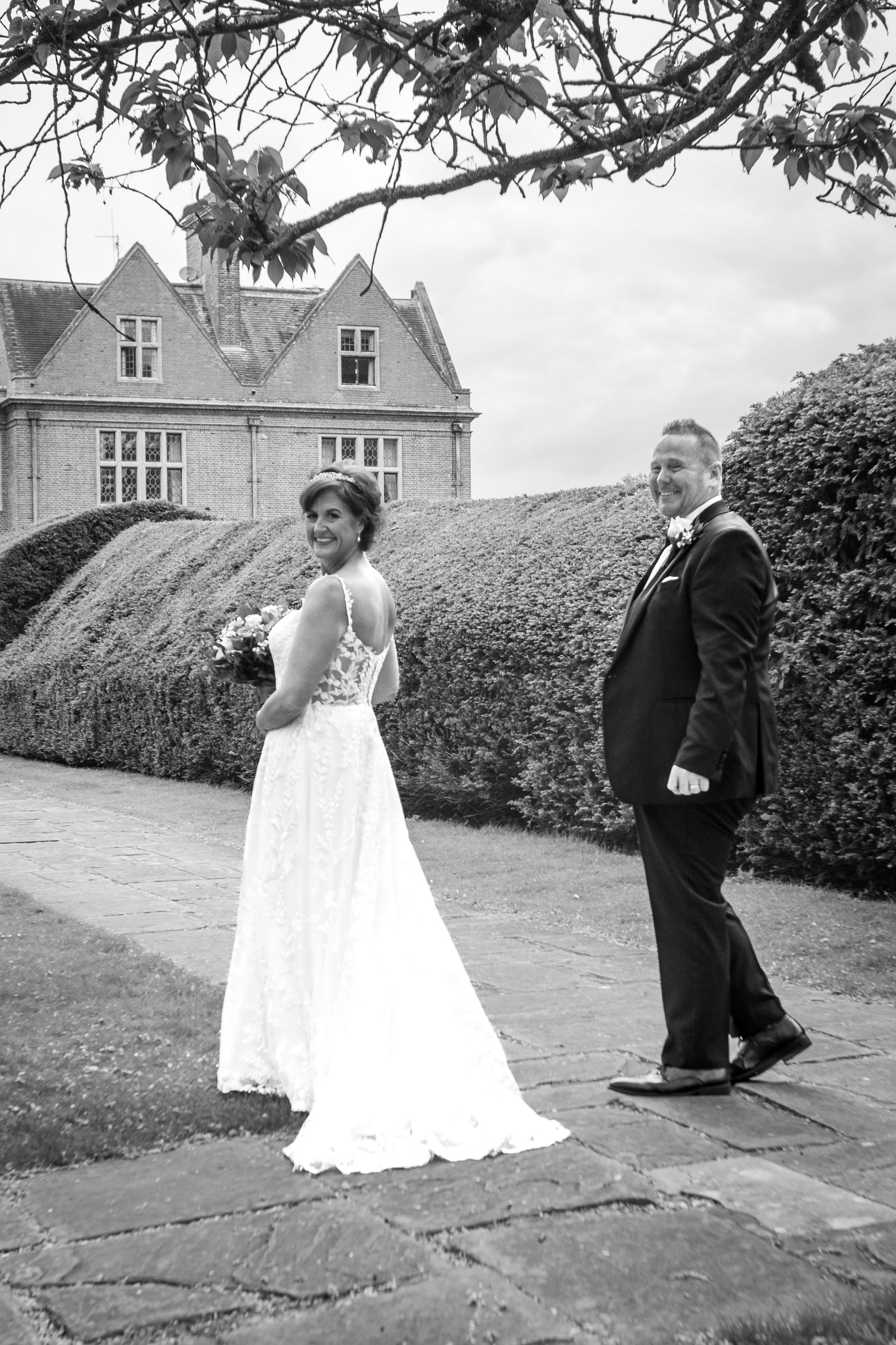 A black and white photo of a bride and groom standing outdoors on a stone path, with the bride holding a bouquet and smiling at the camera while the groom looks at her, in front of a large hedge and an old brick building with multiple gables.