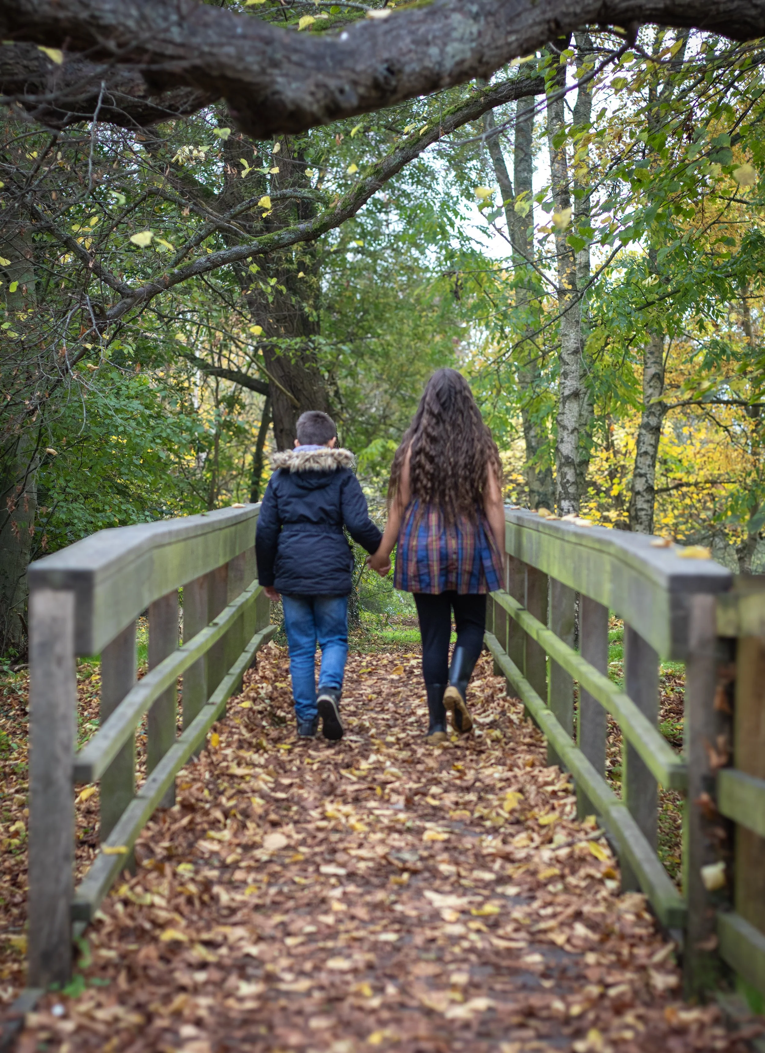 Two children, a boy and a girl, walking hand in hand on a wooded trail with fallen leaves, surrounded by green and autumn-colored trees.
