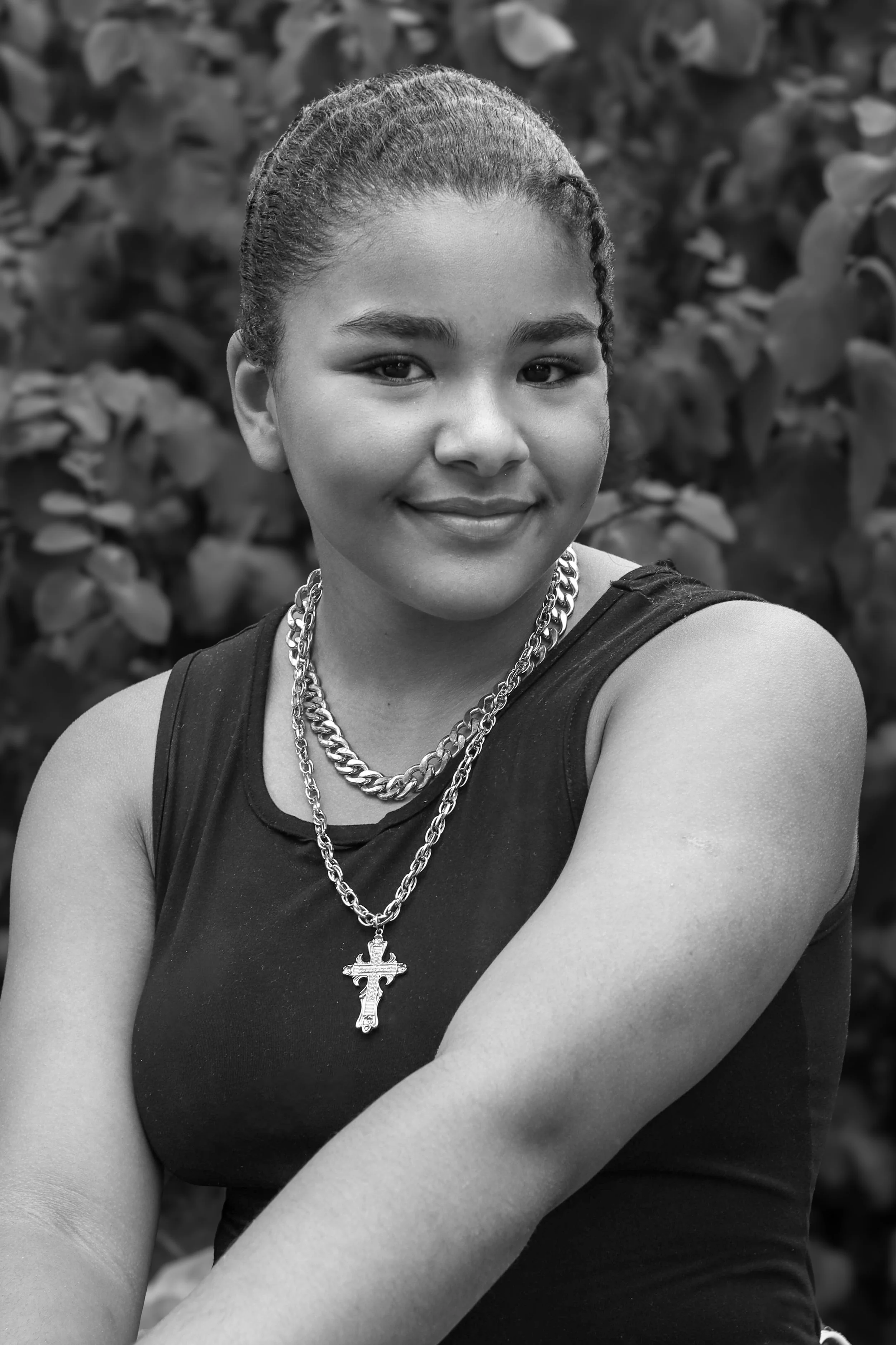 Black and white photograph of a young woman with styled hair, wearing a sleeveless top and layered necklaces, smiling with confident expression, standing in front of leafy background.