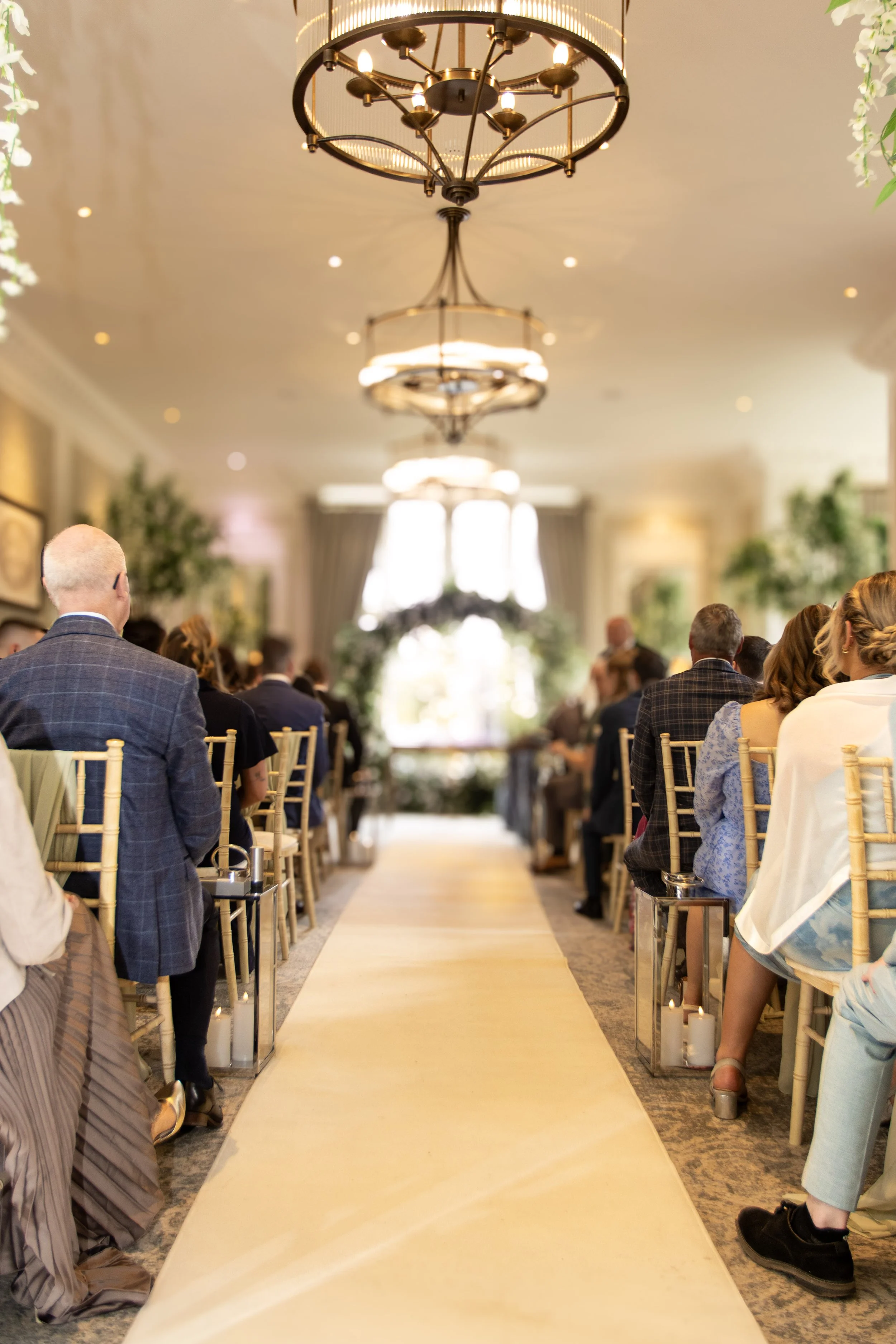 A wedding ceremony in an elegant, well-lit room with guests seated on chairs on both sides of a beige aisle runner. The background features large windows with curtains and greenery, adorned with floral decorations. The ceiling has ornate chandeliers.