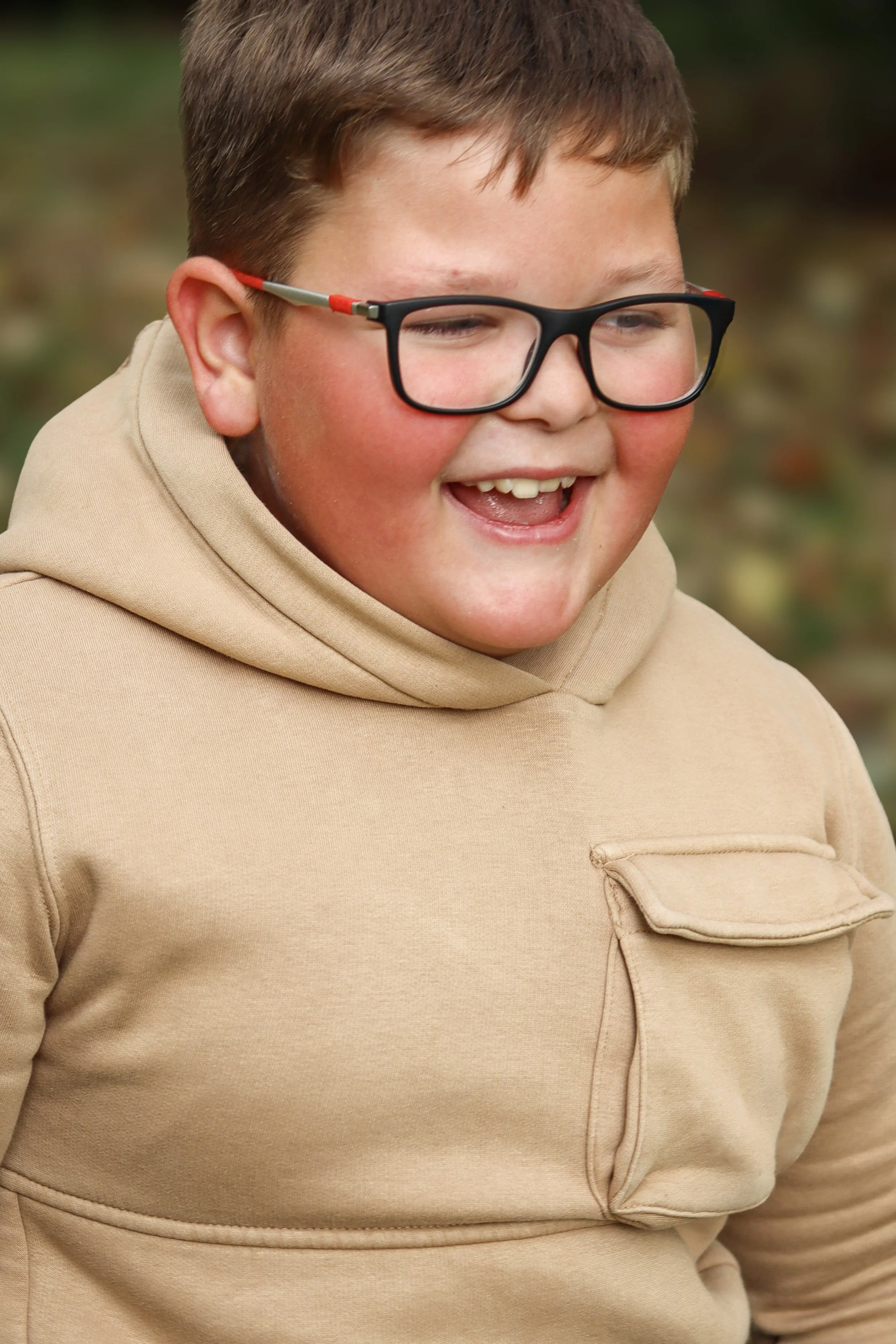 Close-up of a young boy with glasses smiling outdoors, wearing a beige hoodie.