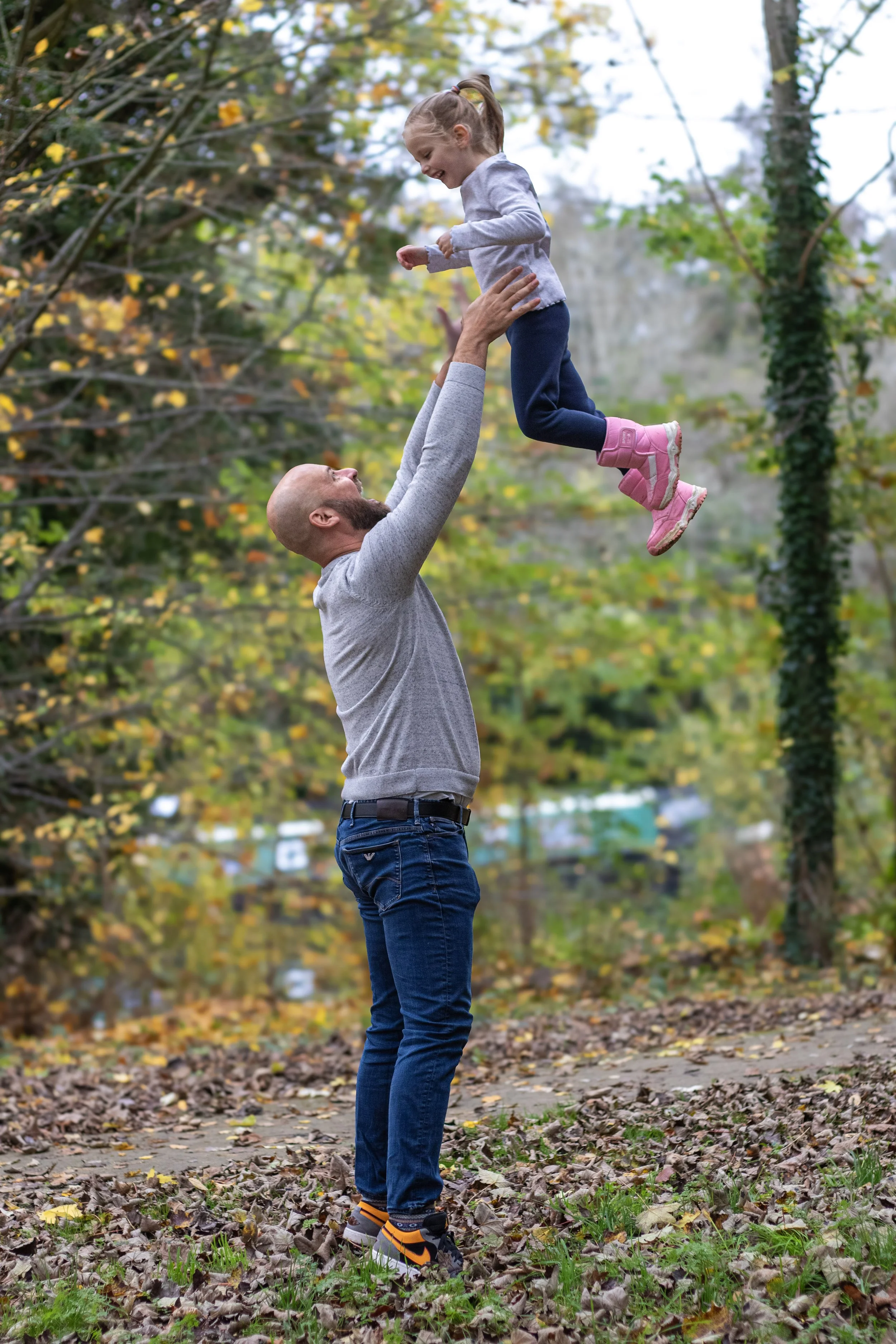 A man is lifting a young girl in the air outdoors during fall, surrounded by trees with yellow and green leaves.