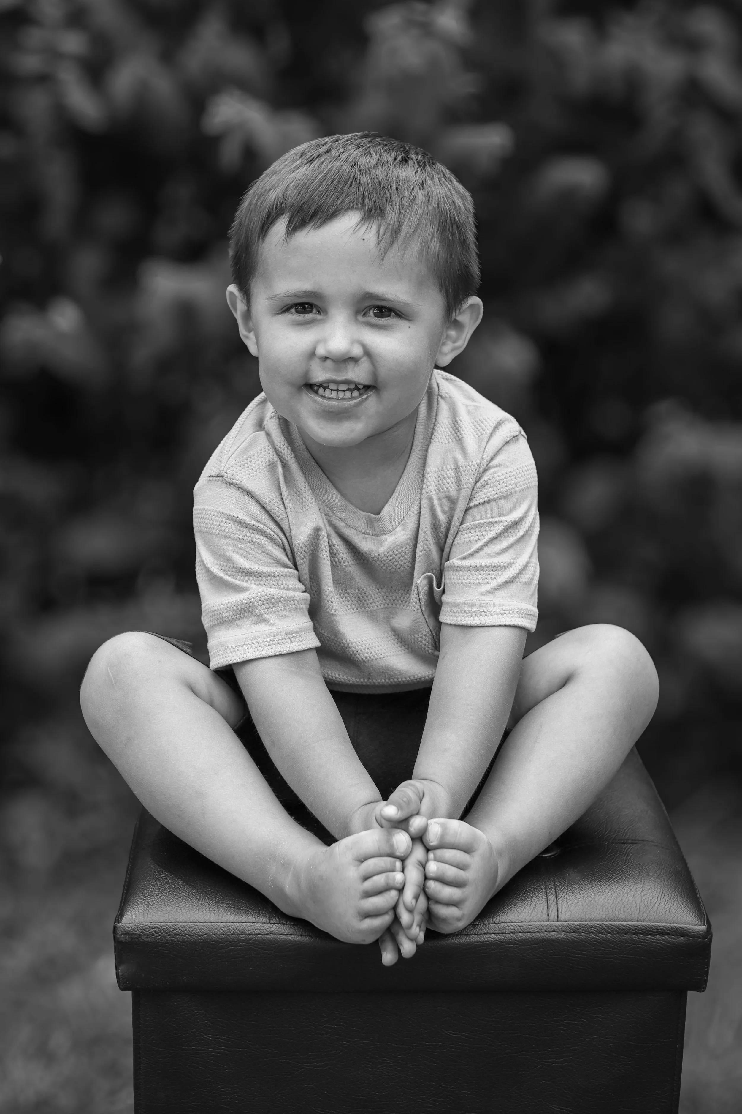 A young boy sitting cross-legged on a padded stool outdoors, smiling at the camera in black and white.