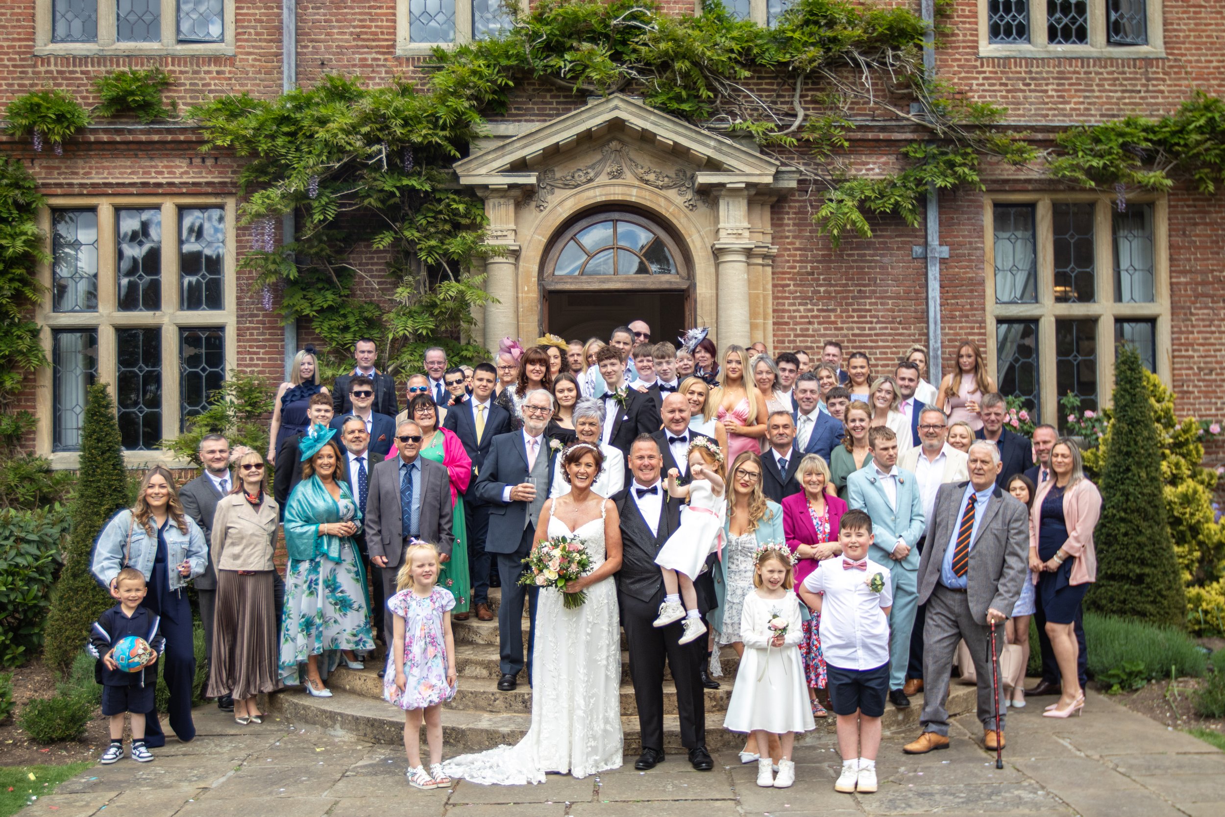 A large wedding party gathering outside a church with brick walls, featuring a bride in a white dress holding a bouquet, a groom in a black tuxedo, and numerous guests dressed in formal attire, including children, women, and men, standing on stone steps and a garden pathway.