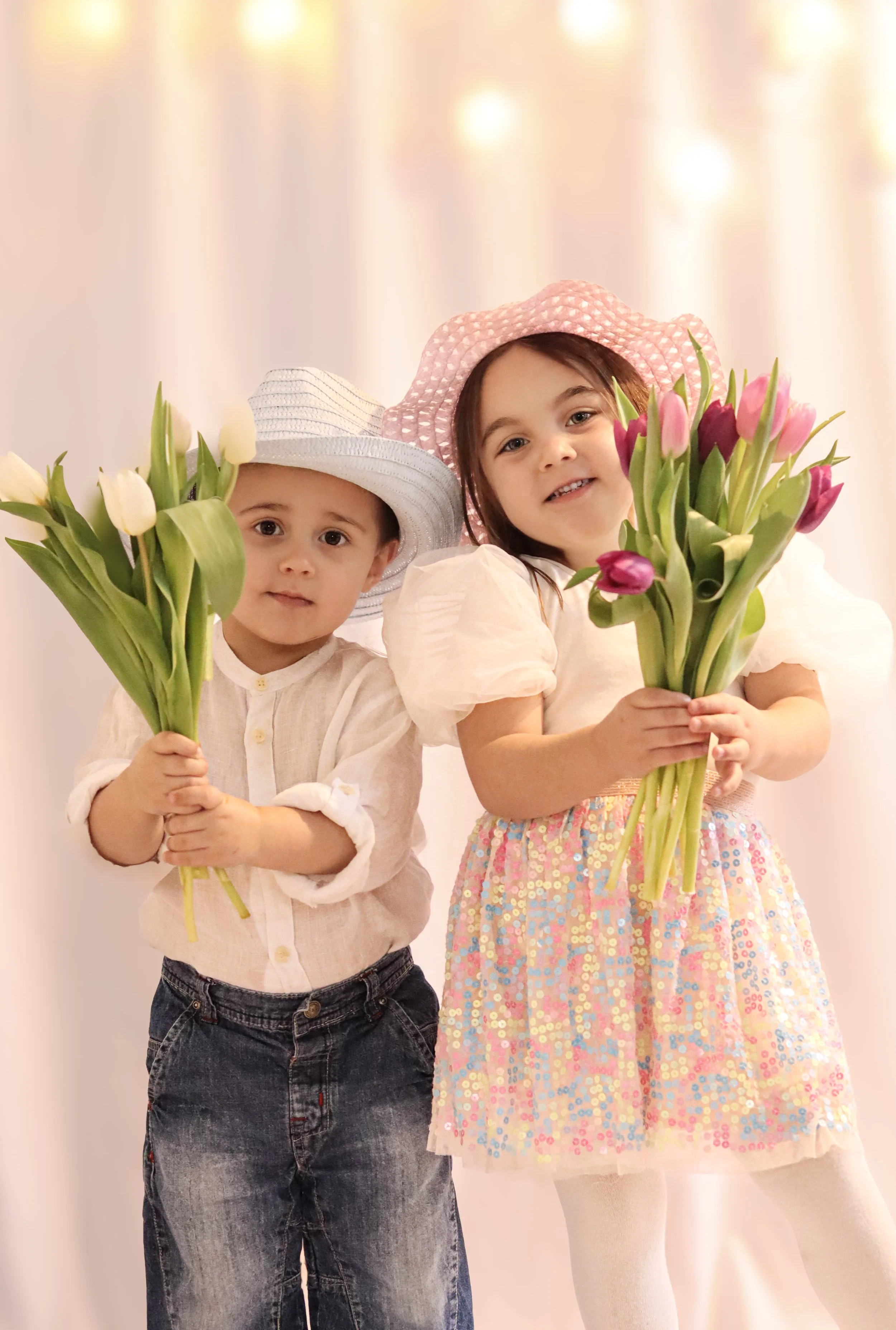 Two children, a boy and a girl, holding bouquets of tulips, wearing pastel outfits and wide-brimmed hats, standing against a soft, glowing background.